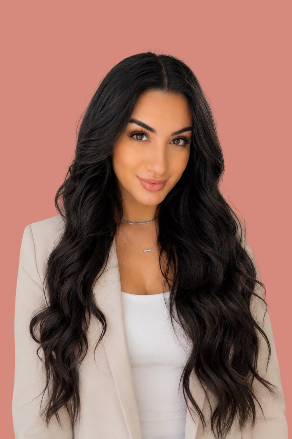 A woman with long, dark, wavy hair and light makeup posing against a pink background, wearing a white top, beige blazer, and layered necklaces.