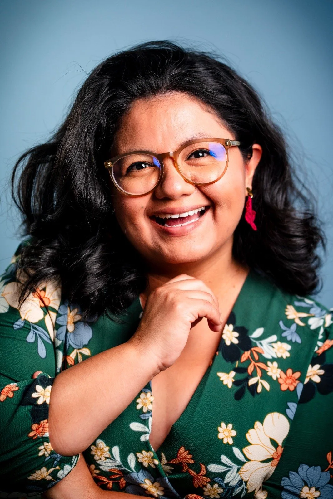 Close-up of a smiling woman with glasses, dark curly hair, wearing a floral green top and pink earrings, against a blue background.
