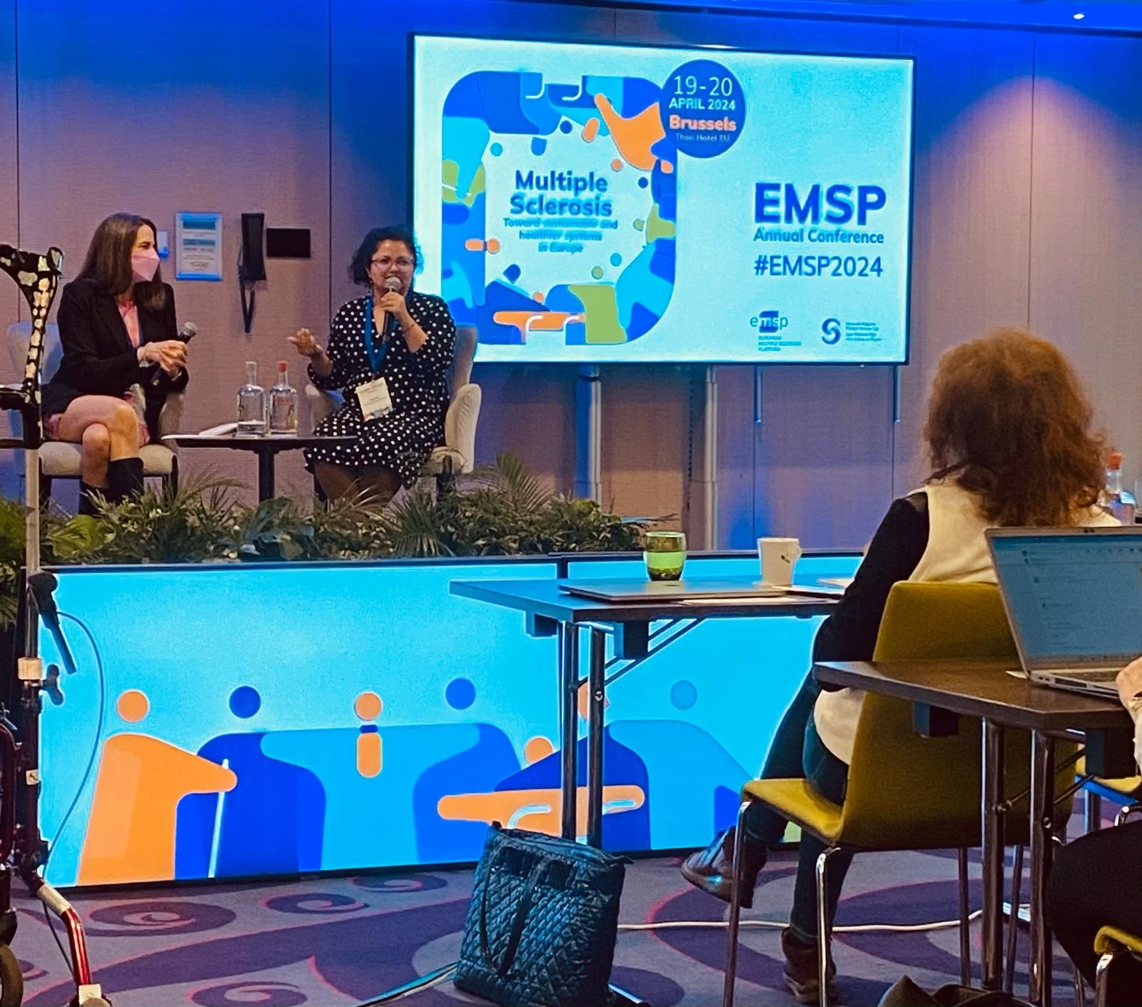 At a conference, two women sit on a stage engaging in a discussion with microphones in hand, while an audience member takes notes. A large screen behind them displays information about a Multiple Sclerosis conference scheduled for April 19-20, 2024, in Brussels, with various logos and branding elements.