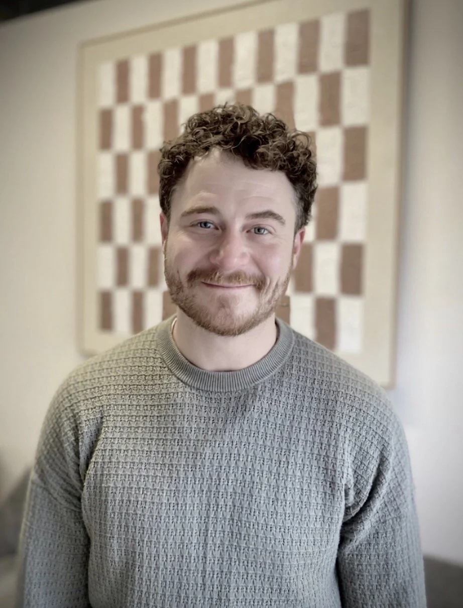 A young man with curly brown hair and a beard, smiling, wearing a textured green sweater, standing indoors in front of a patterned wall.