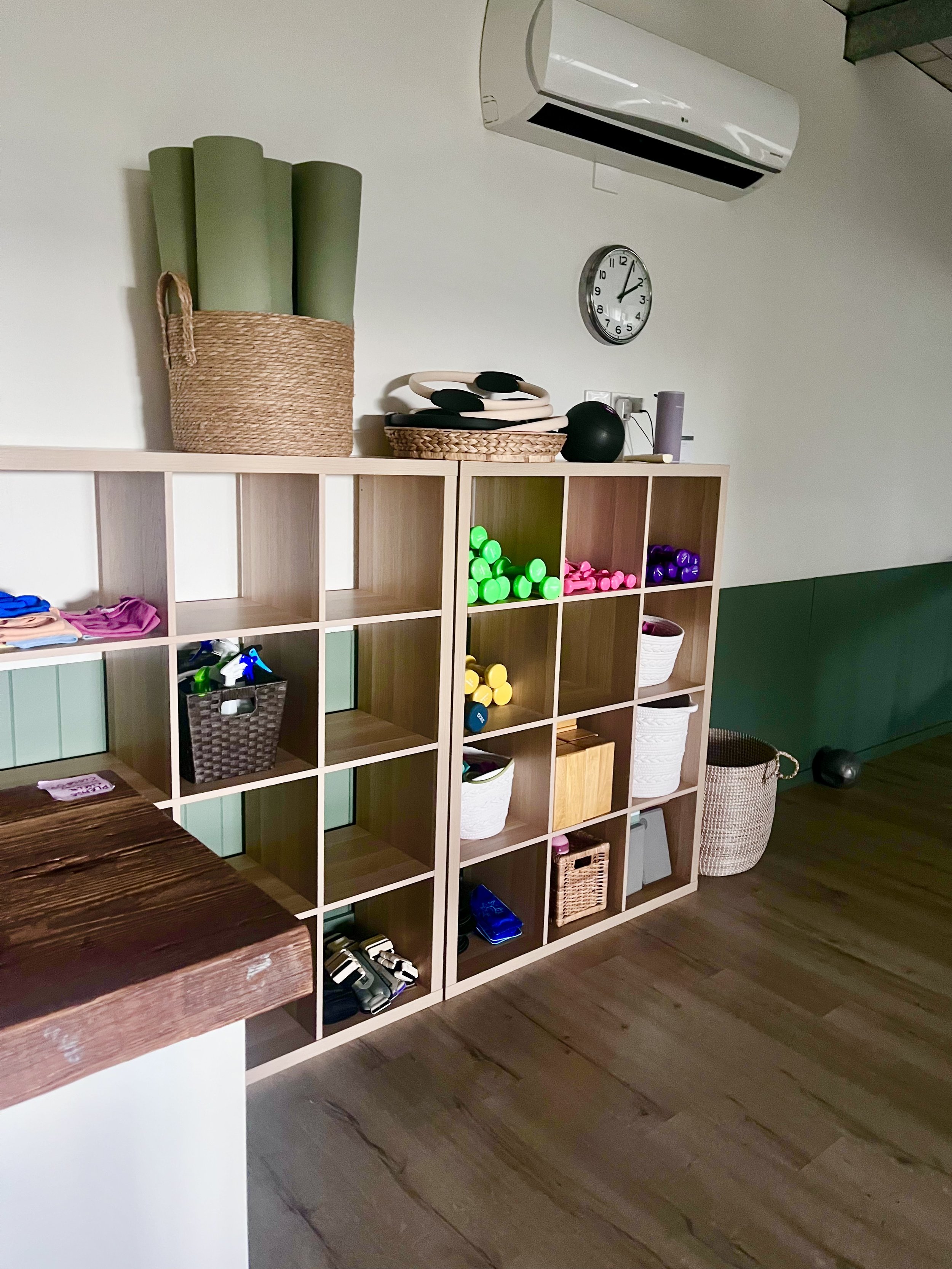 A wooden shelf unit holds various fitness and wellness items, including foam rollers, resistance bands, and dumbbells. There is an open floor plan where yoga and Pilates classes operate.