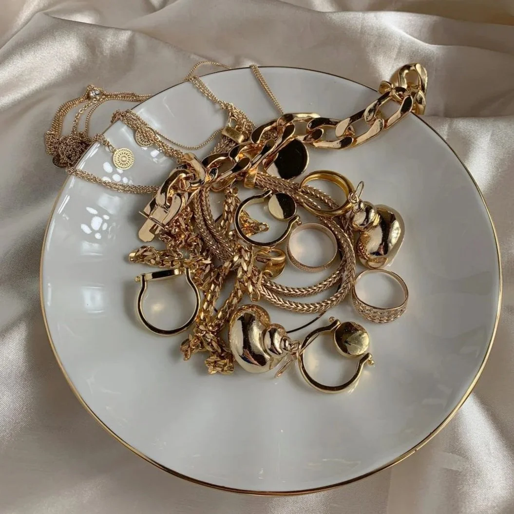 Une assiette blanche avec de nombreux bijoux en or, y compris des chaînes, des bagues et des boucles d'oreilles, posée sur un tissu soyeux beige.