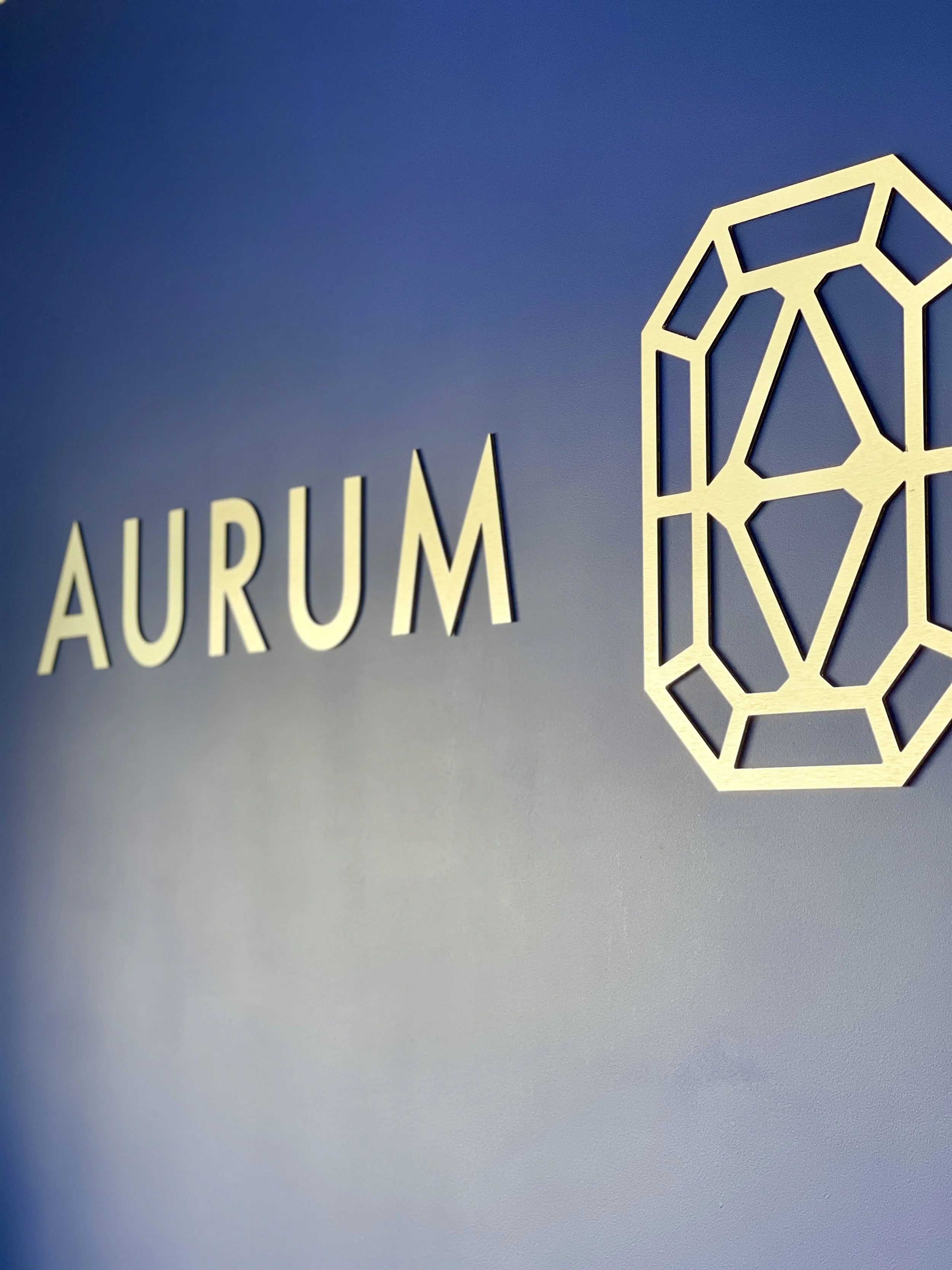 Le mot 'AURUM' écrit en lettres majuscules devant un mur bleu avec un symbole géométrique en fil de métal à côté.