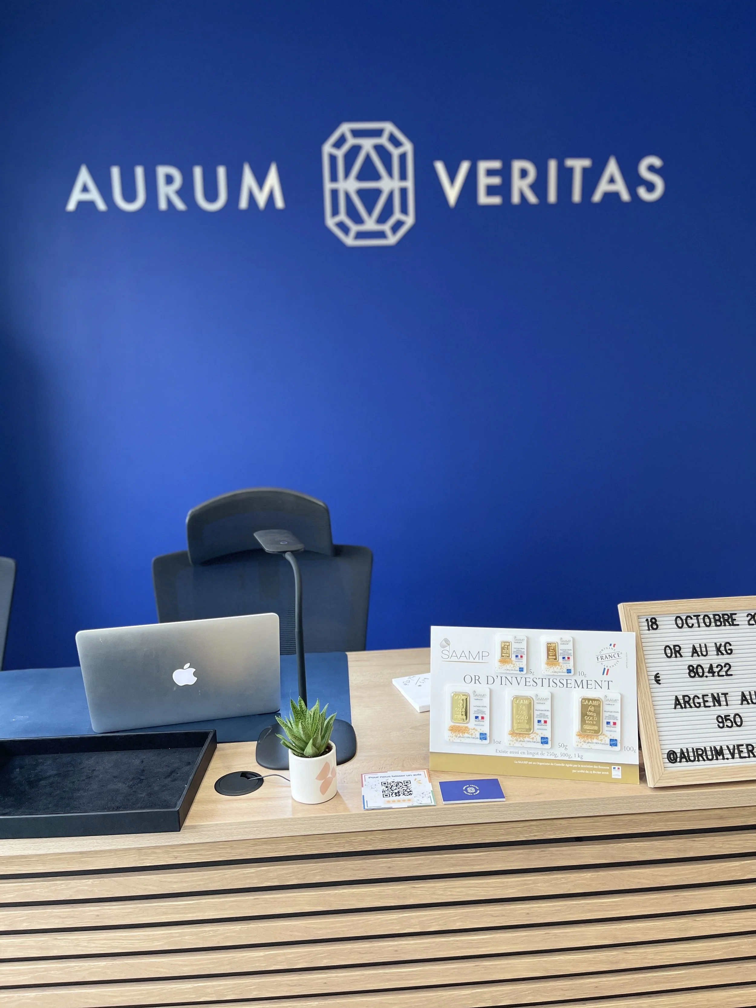 Un bureau avec un ordinateur portable Apple, un écran, des plaques d'or, des notices et un tableau blanc avec des prix pour l'or, l'argent et le platine.