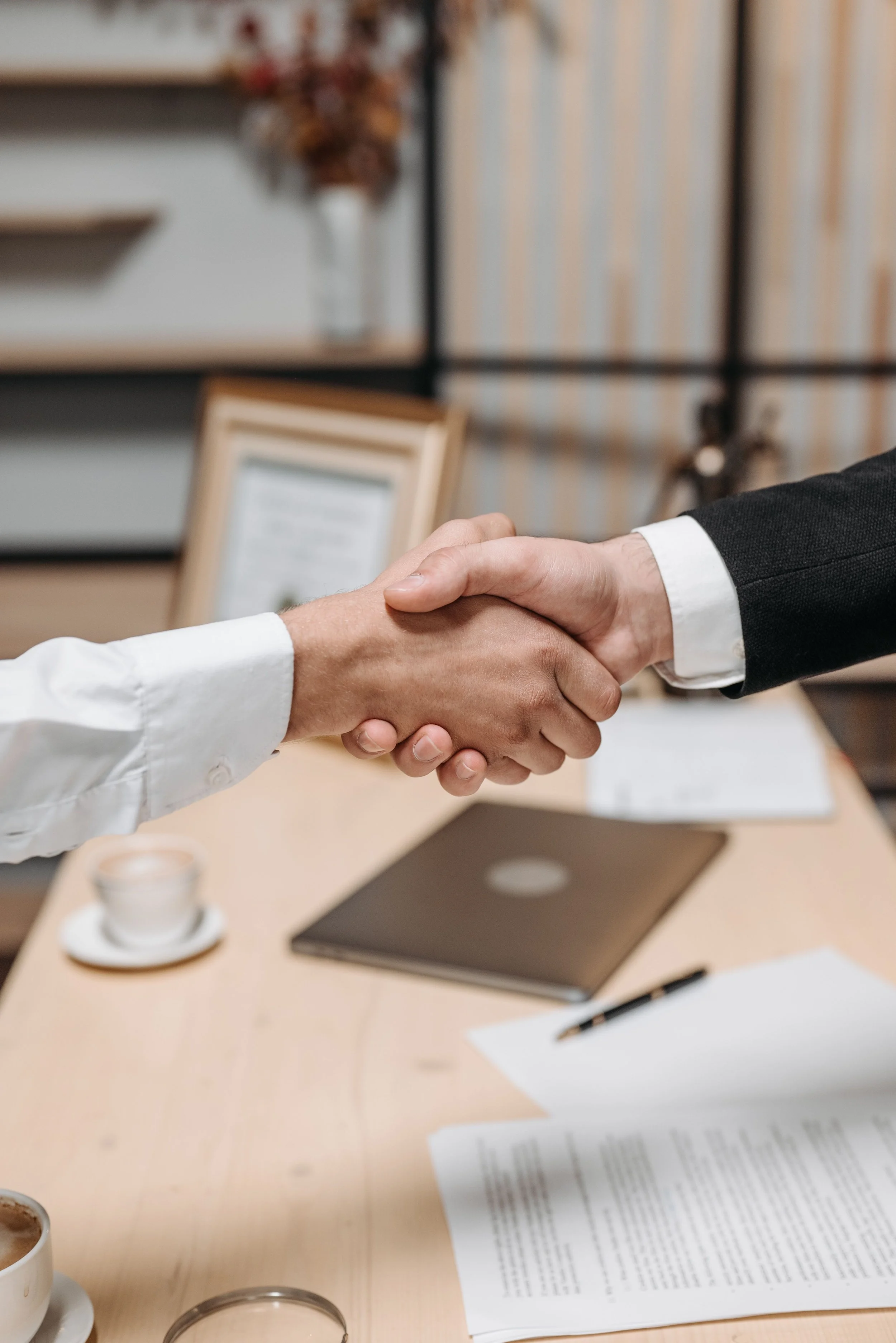 Deux personnes se serrent la main lors d'une réunion de travail, avec un bureau, un ordinateur portable, des documents et une tasse de café sur la table.