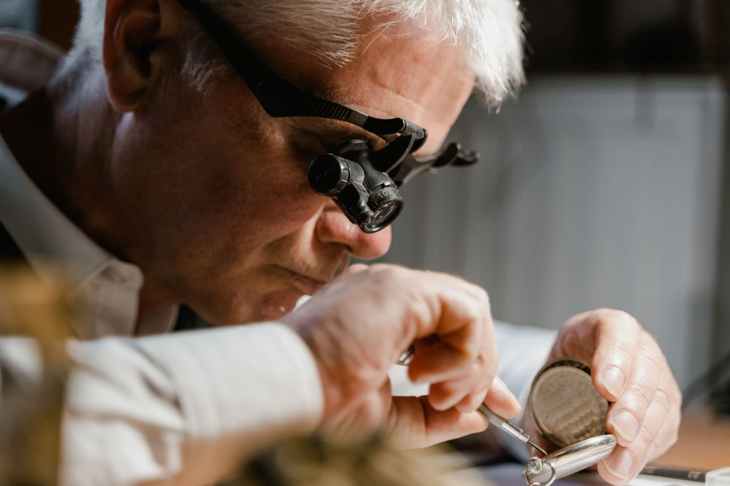 Un homme âgé avec des lunettes de loupe se concentre sur un objet qu'il tient dans ses mains, en utilisant une loupe pour examiner de près, avec un fond flou.