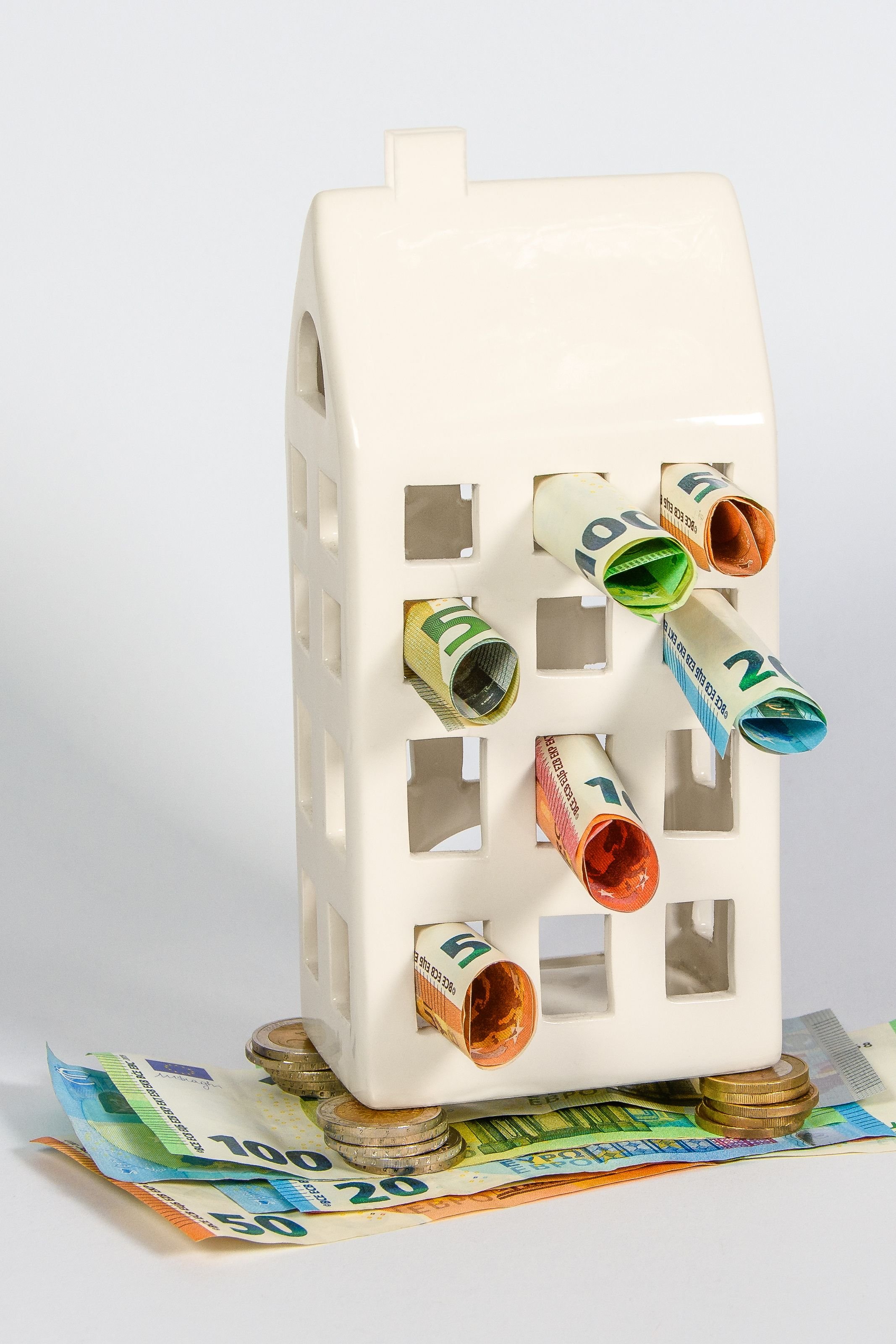 Une petite maison blanche en plastique avec des rouleaux de billets de banque et des pièces de monnaie empilées dessous et autour, représentant une idée de richesse ou d'épargne.