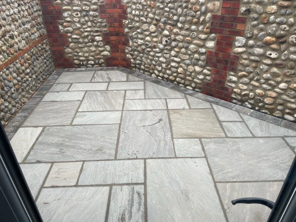View of a small outdoor corner patio with light gray stone paving and walls made of small rounded river rocks and red brick accents.
