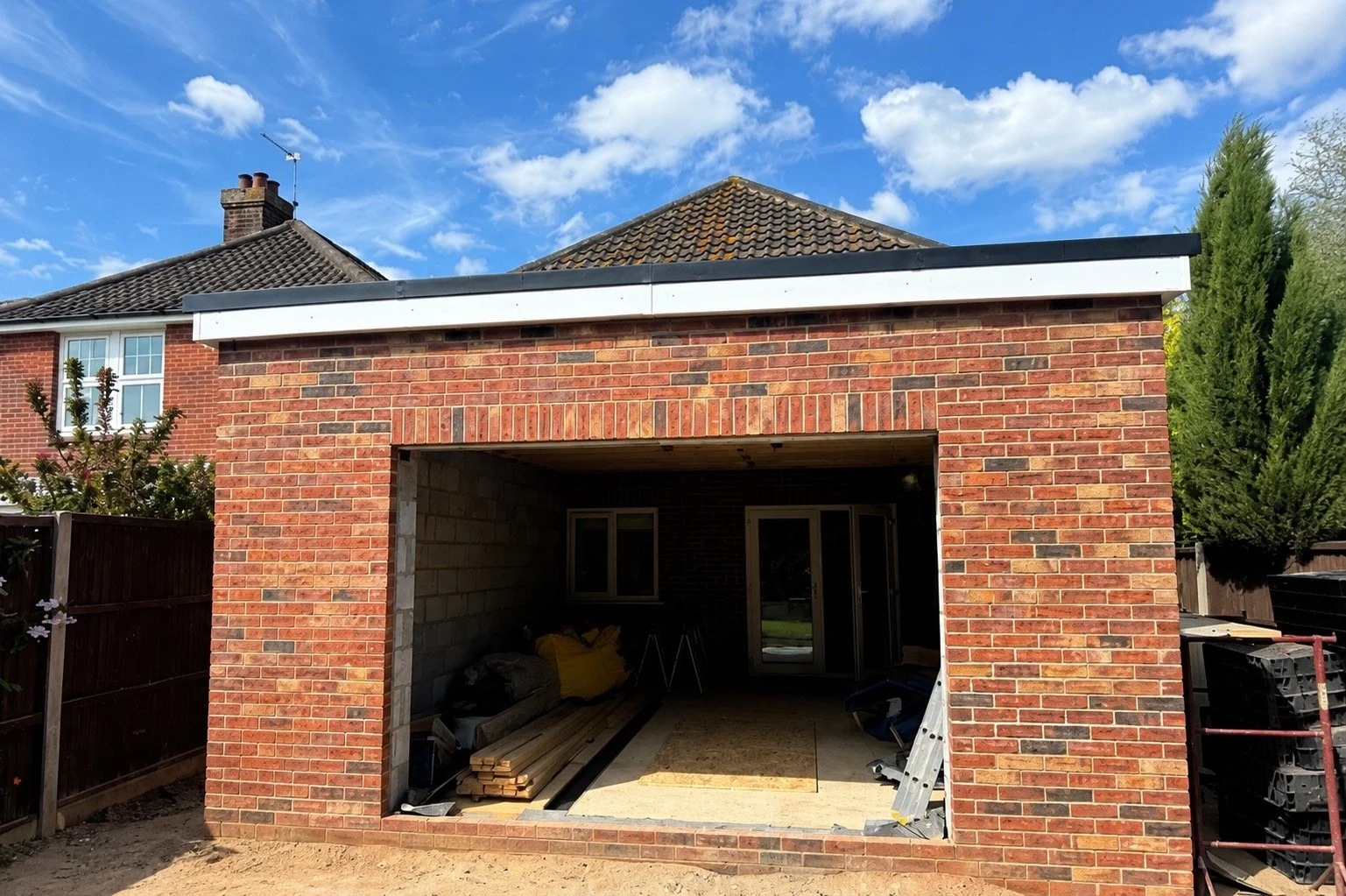 A brick garage being constructed behind a red brick house, with construction materials and equipment inside and a clear blue sky above.