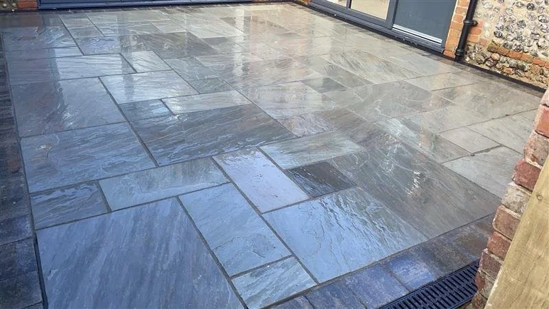 Wet, grey stone patio with a drainage grate near the brick wall.