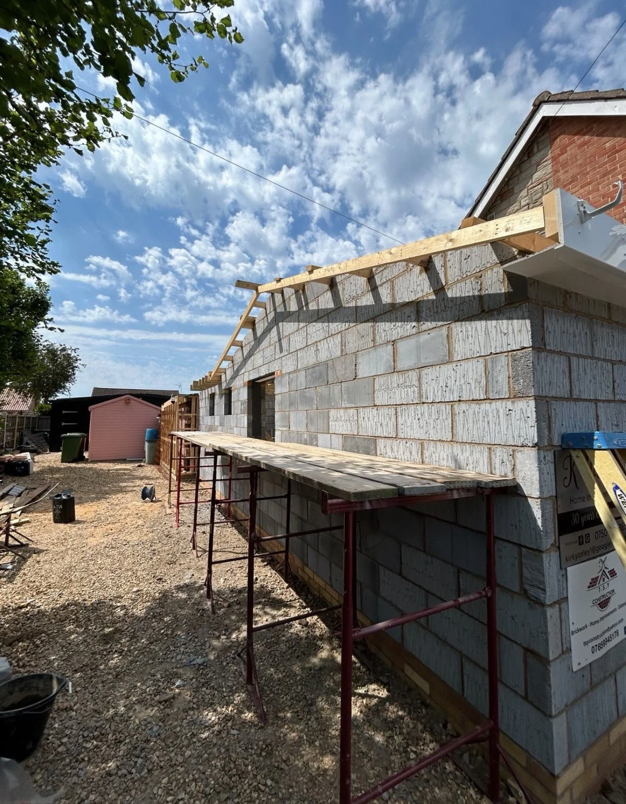 Construction site with an unfinished brick house, scaffolding, and construction materials under a partly cloudy sky.