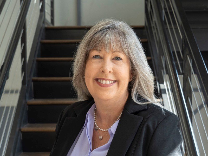 A smiling woman with gray hair, wearing a black blazer and white shirt, standing on a staircase in an indoor setting.