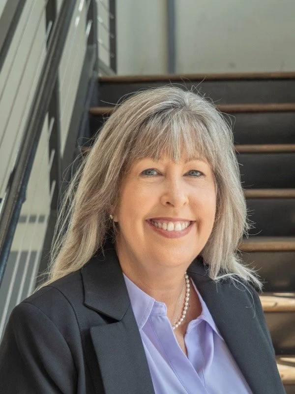 A woman with shoulder-length gray hair, wearing a dark blazer, light purple shirt, and pearl necklace, smiling outdoors near a staircase.