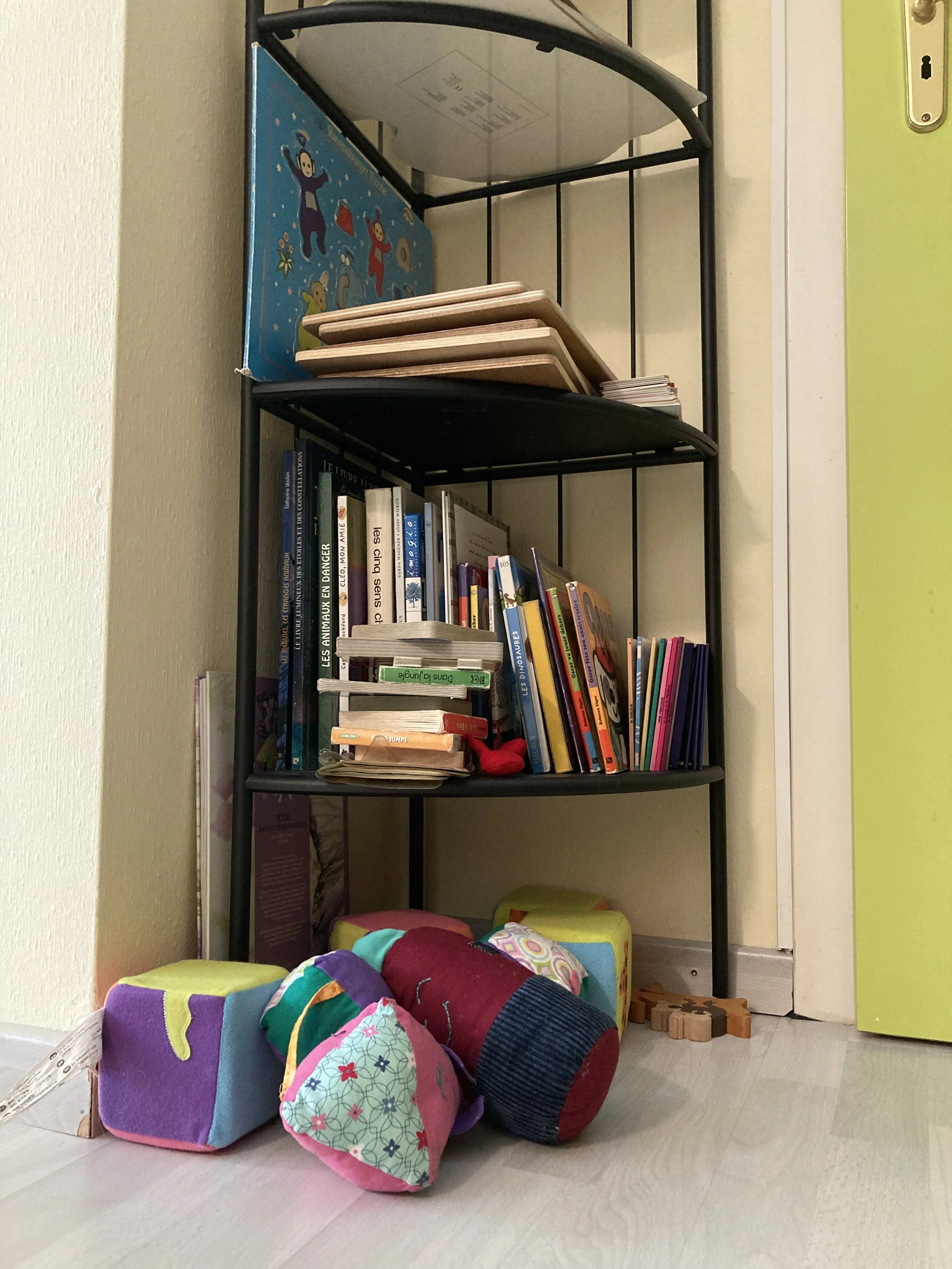 Une étagère noire contenant des livres, des cahiers, et des livres pour enfants. Au sol, des jouets en tissus multicolores.