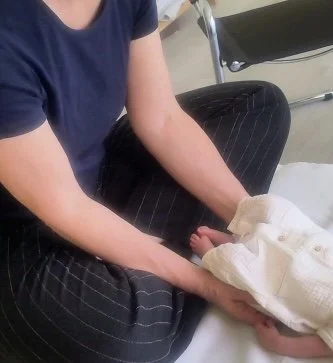 Une personne assise portant un t-shirt bleu et des pantalons noirs à rayures, tenant un bébé enveloppé dans une couverture blanche.