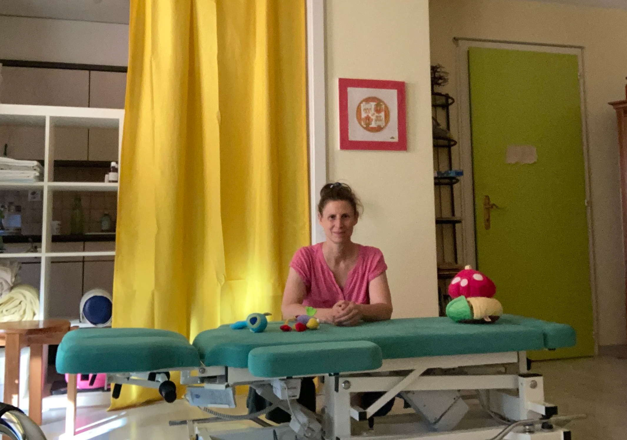 Une femme assise à une table avec des jouets en peluche colorés devant elle, dans une pièce avec un rideau jaune, un mur avec cadre et de l’équipement médical.