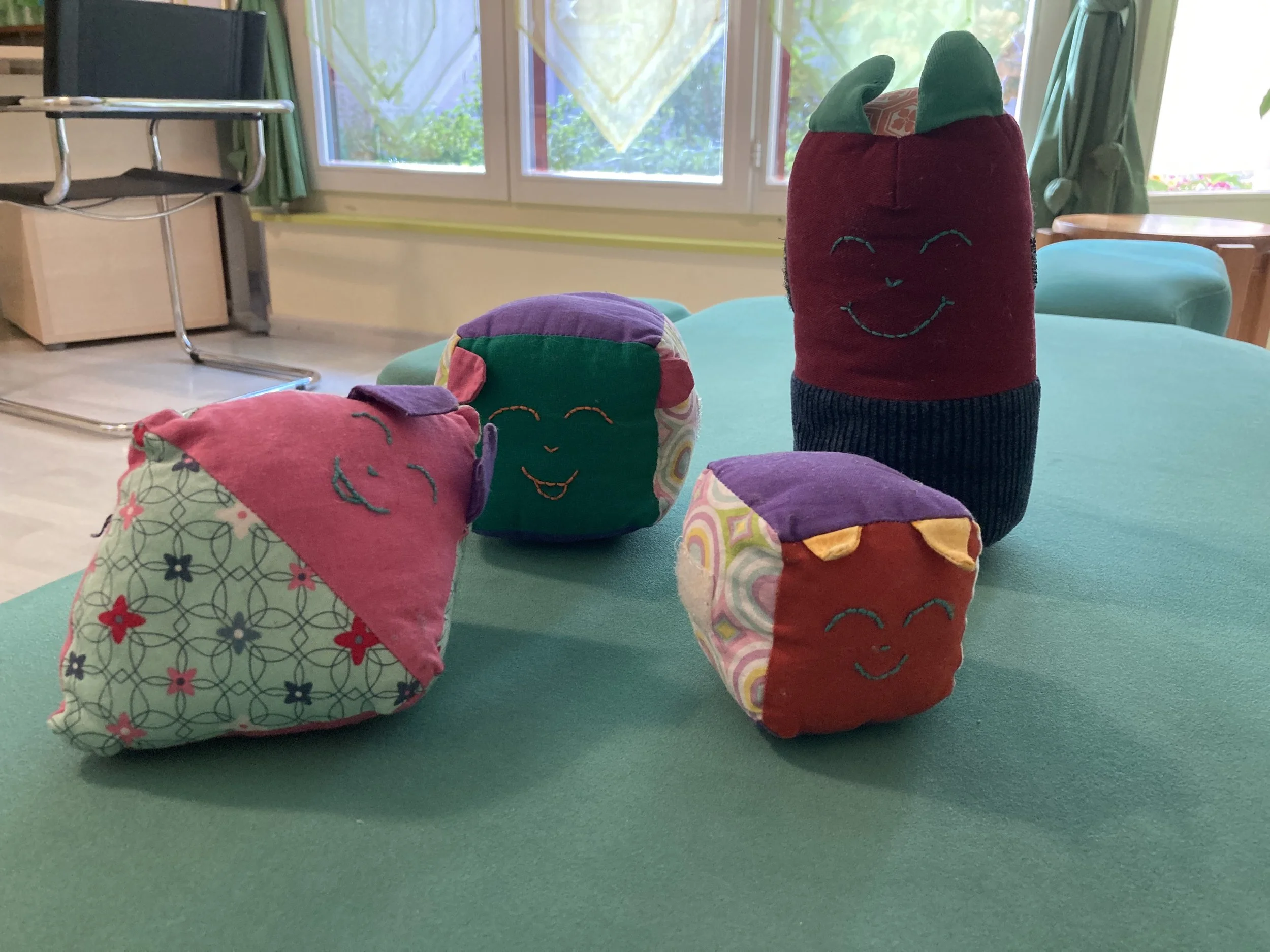 Une collection de personnages en tissu colorés et souriants, disposés sur une table verte dans une pièce lumineuse avec fenêtres et plantes à l'extérieur.