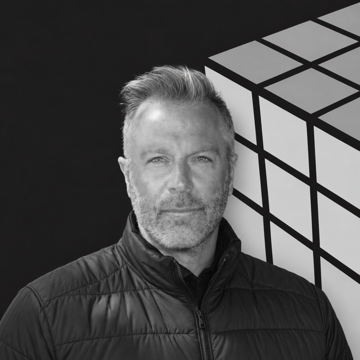 Black and white portrait of a middle-aged man with short, slicked-back hair and a beard, wearing a black zip-up jacket, standing in front of a black background with a partial view of a large geometric shape.
