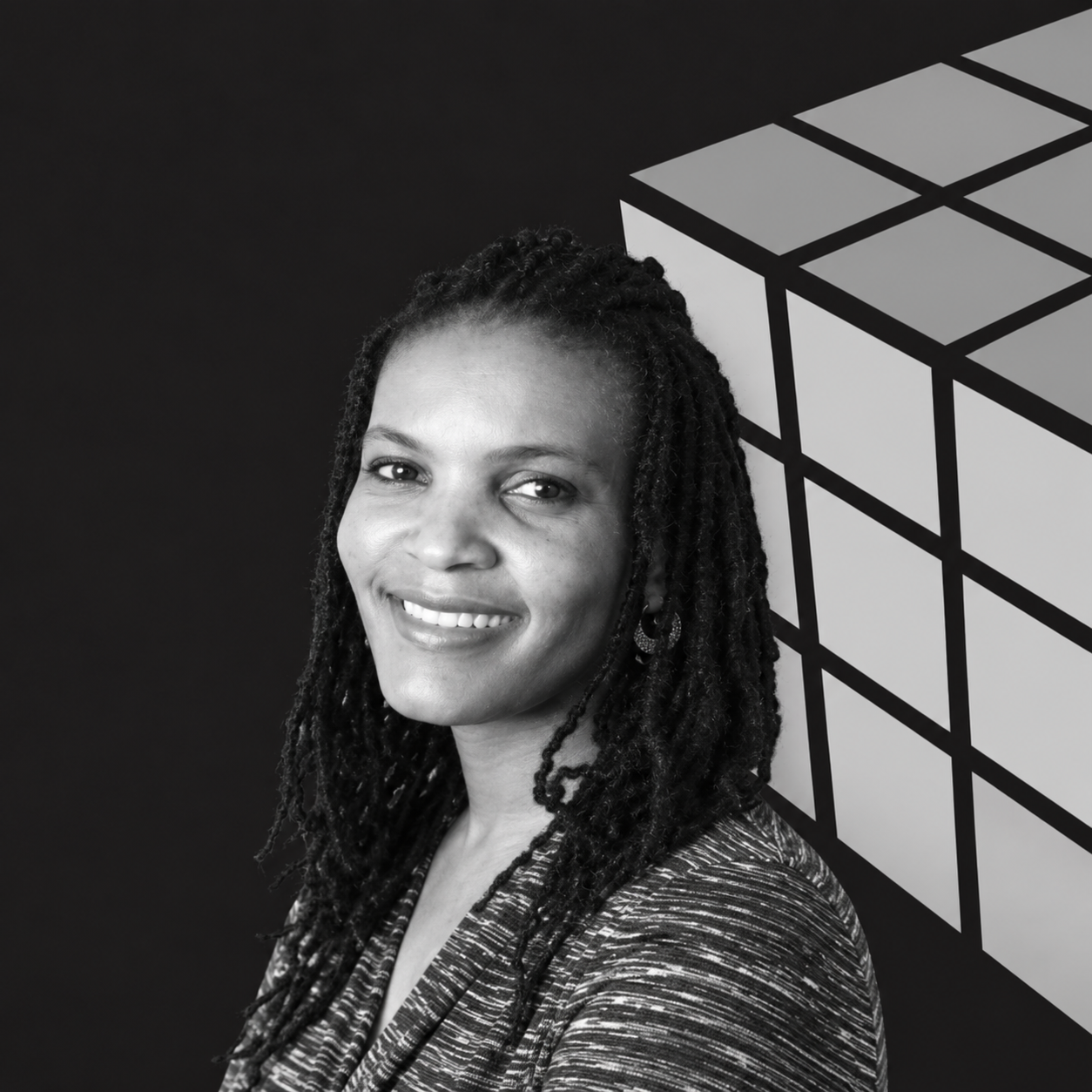 Black and white photo of a smiling woman with dreadlocks standing in front of a geometric pattern background.