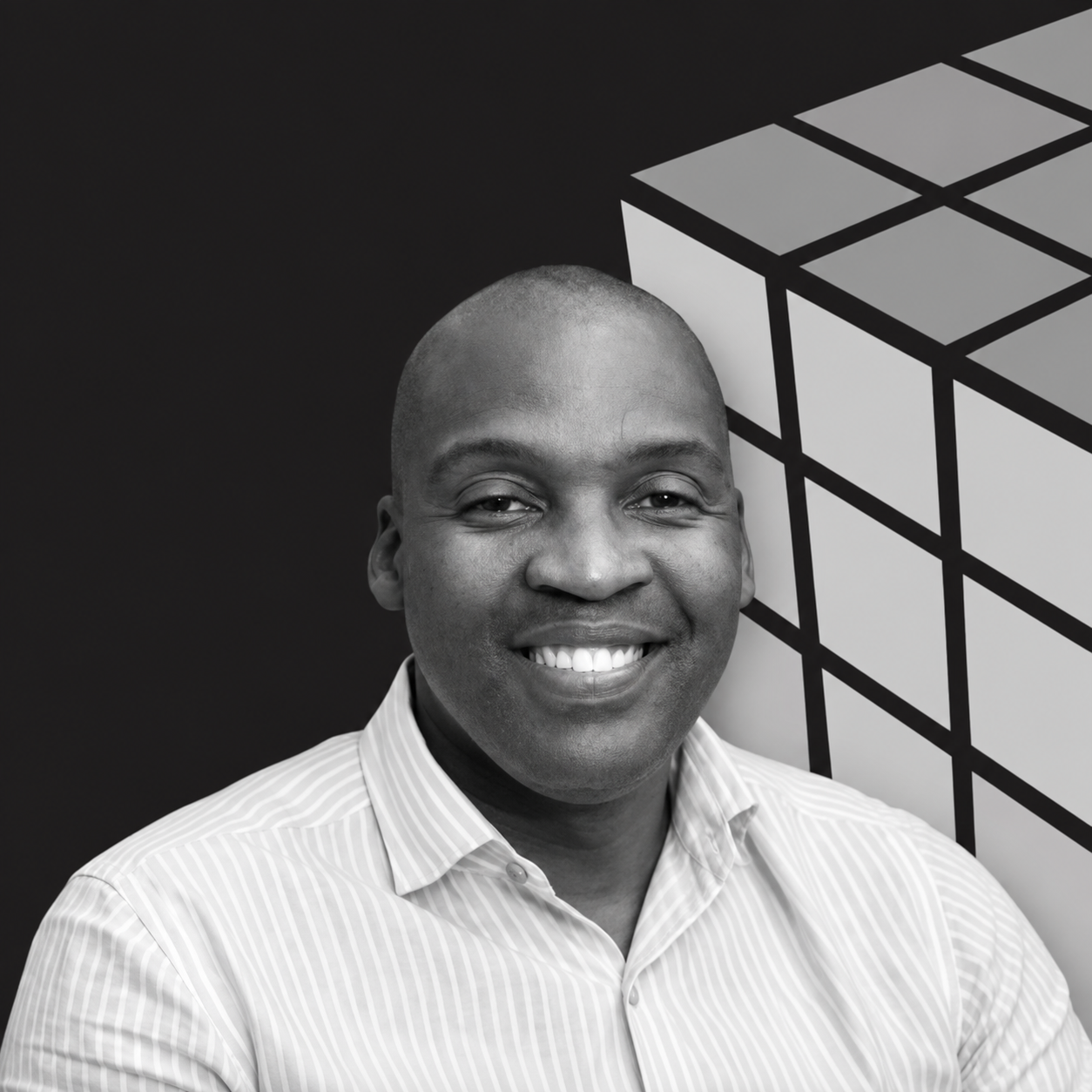 Black and white photo of smiling man wearing a striped button-up shirt, standing in front of a geometric background.
