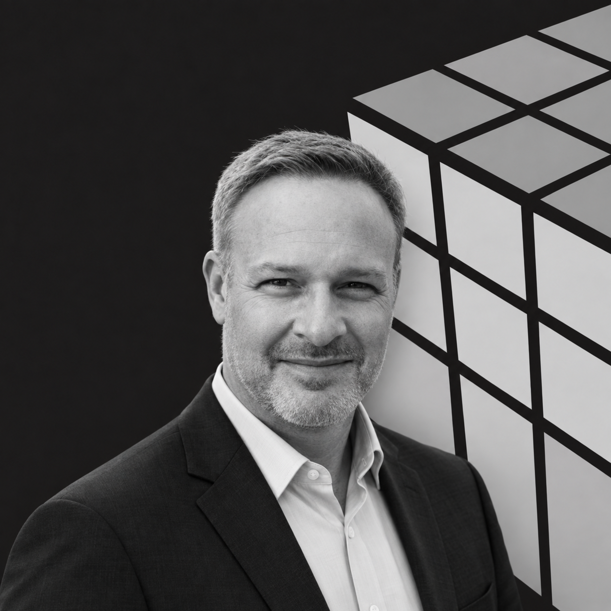 Black and white portrait of a middle-aged man with short hair and light stubble, wearing a suit and white shirt, standing in front of a geometric artwork.