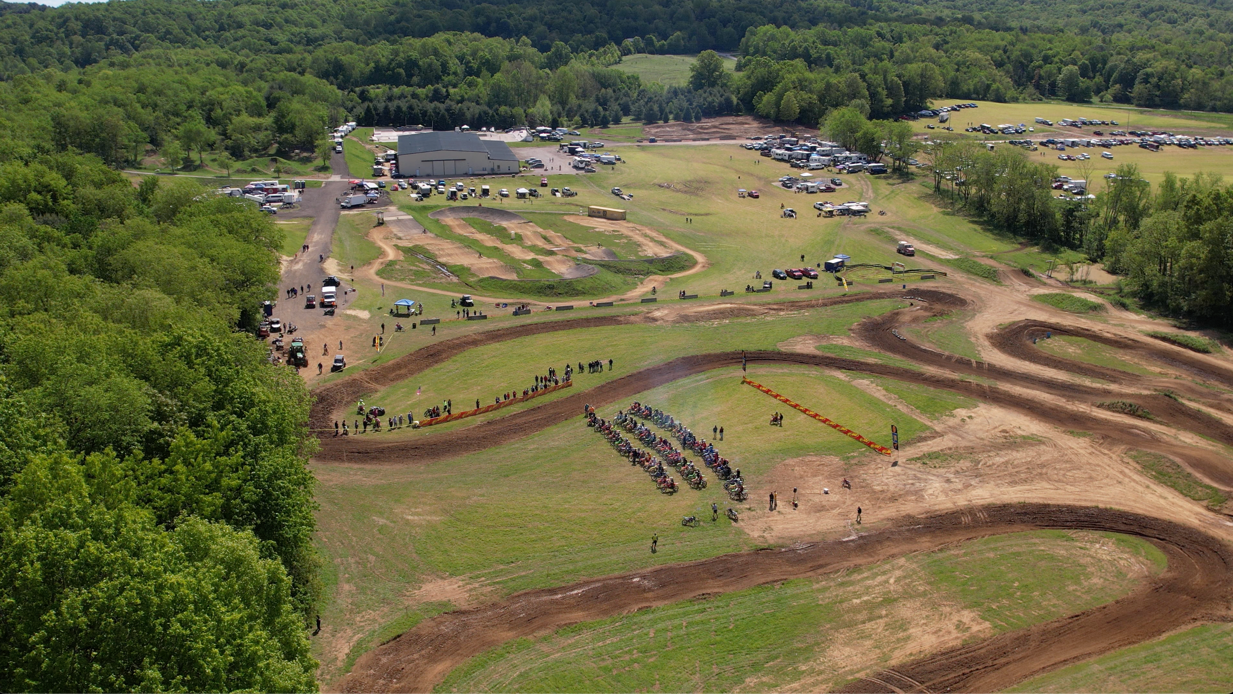 Overhead view of a motocross track with dirt trails, jumps, and turns, surrounded by green trees, and a parking lot with vehicles in the distance.
