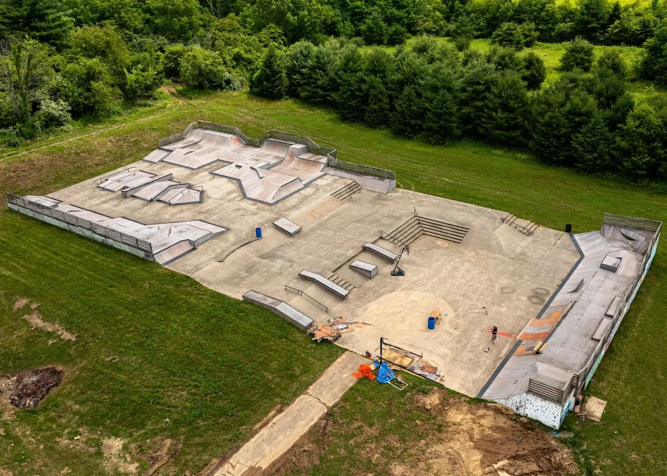 An outdoor skatepark with various ramps, rails, and platforms, surrounded by grassy areas and trees in the background.