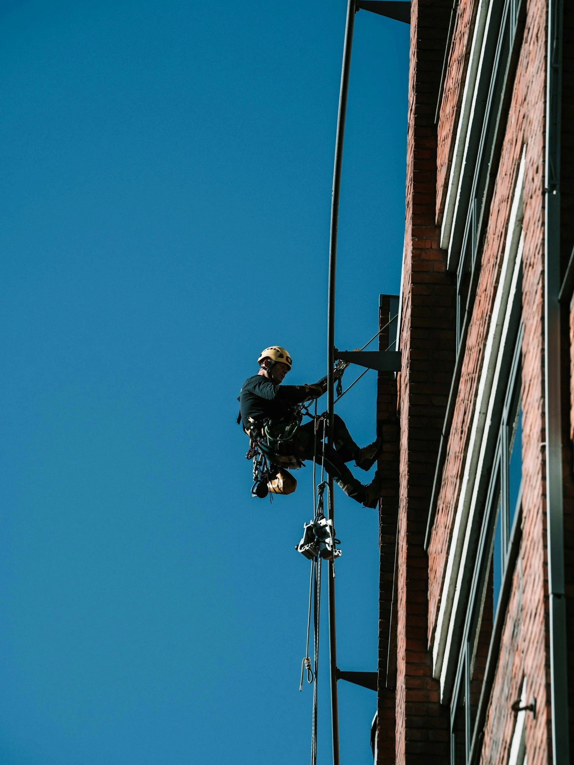 A worker wearing a safety helmet and harness is cleaning windows on a tall brick building using a rope access system.