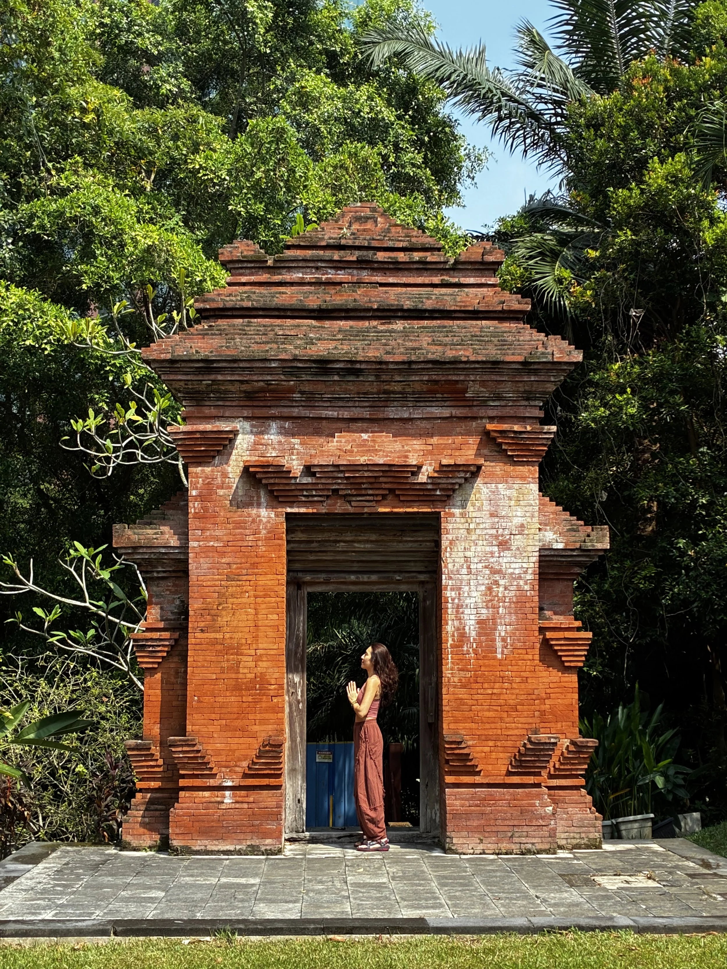 A woman in a yoga pose standing under a small brick structure surrounded by lush green trees.