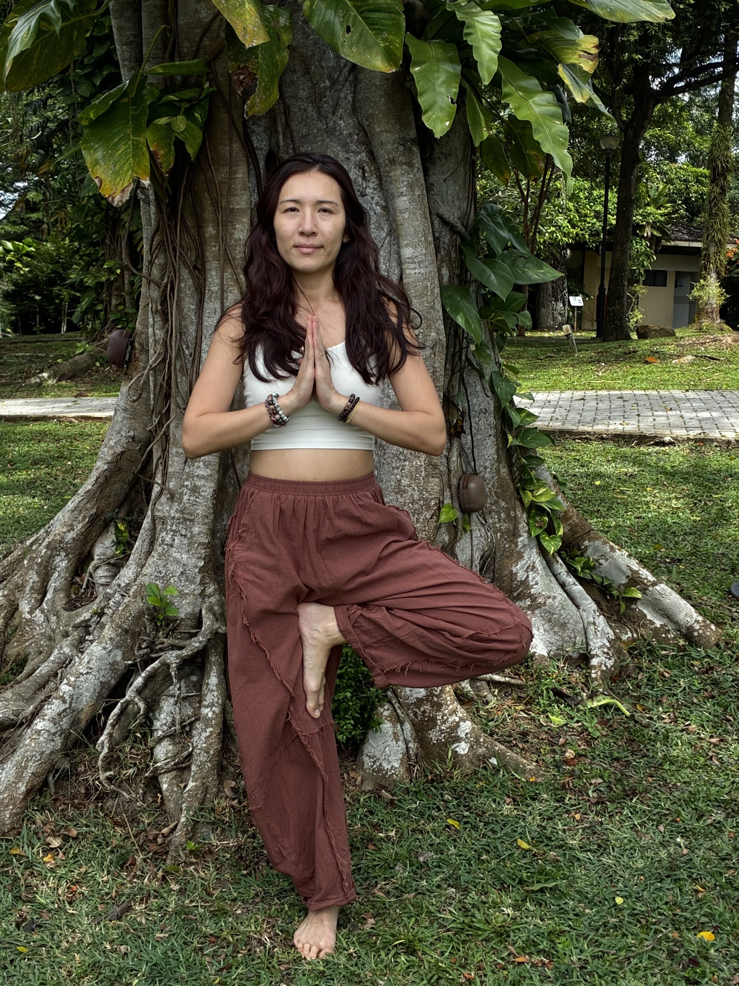 A woman practicing yoga outdoors, standing in tree pose with one foot pressed against the inner thigh of her other leg, hands in prayer position, next to a large tree with green leaves.