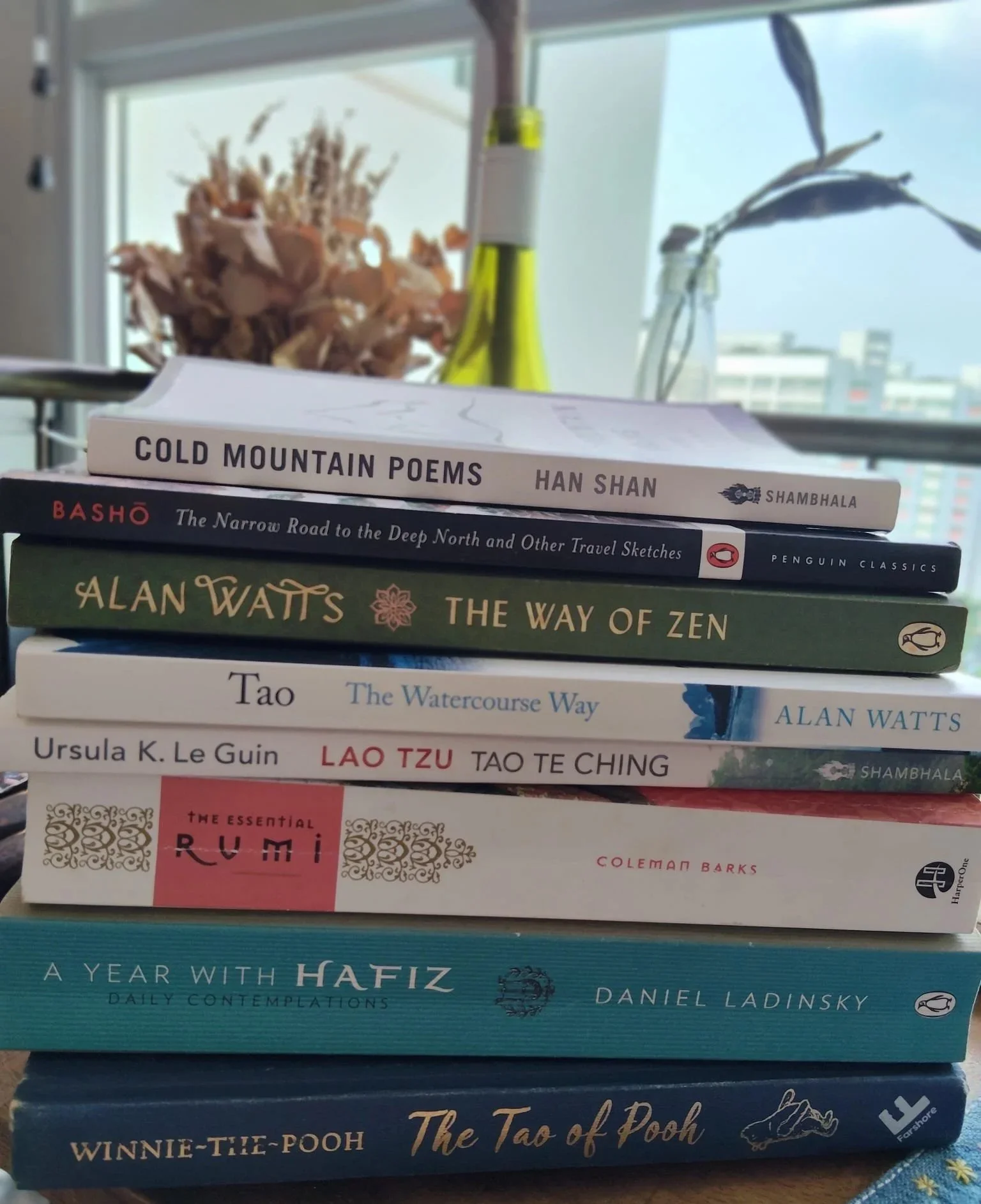 Stack of books with titles related to Asian philosophy, travel, and culture, placed on a table near a window with decorative dried flowers and a lemon-scented candle.