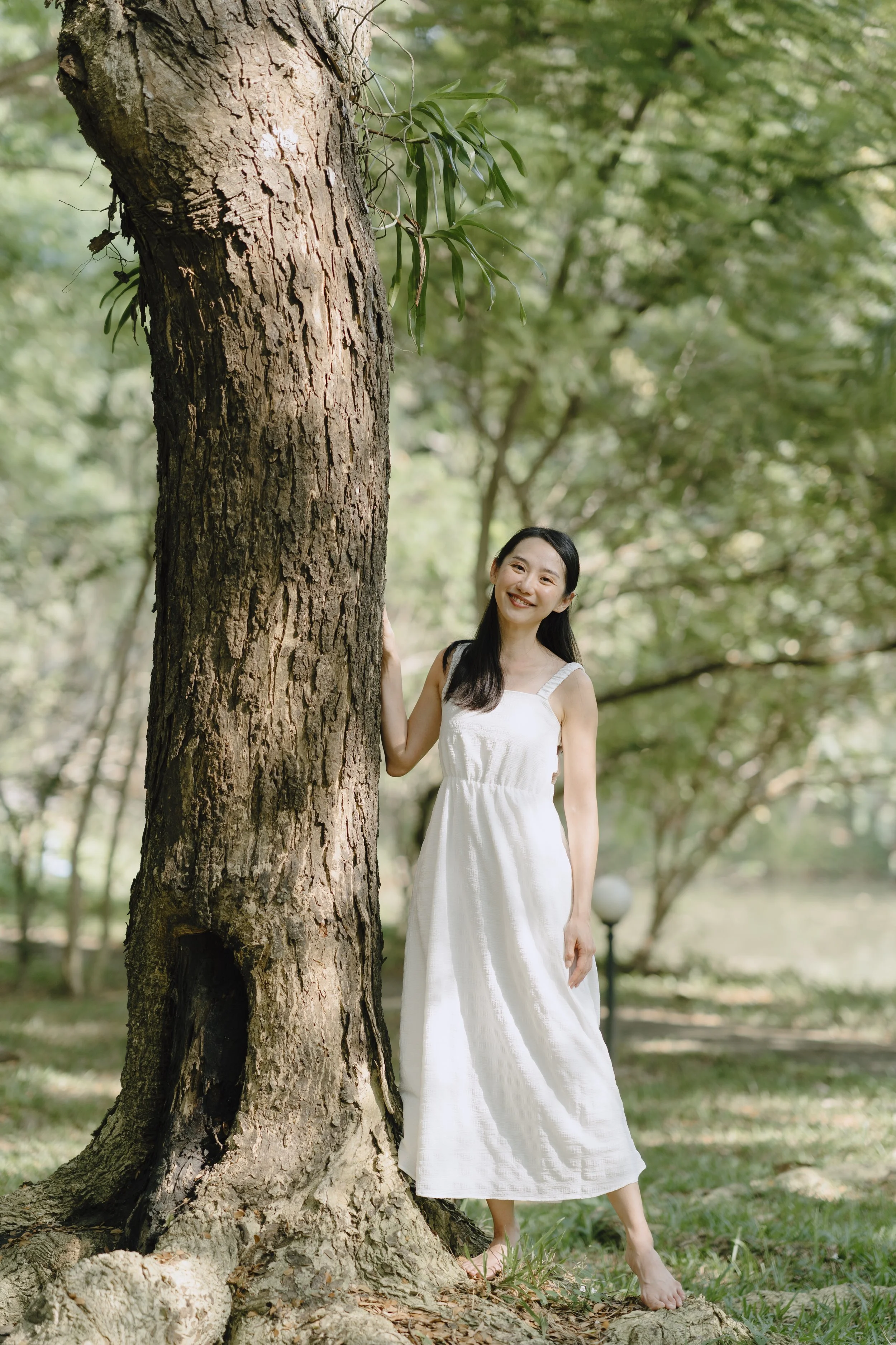 A young woman with long dark hair in a white sundress standing outdoors behind a large tree with green leaves, smiling at the camera.