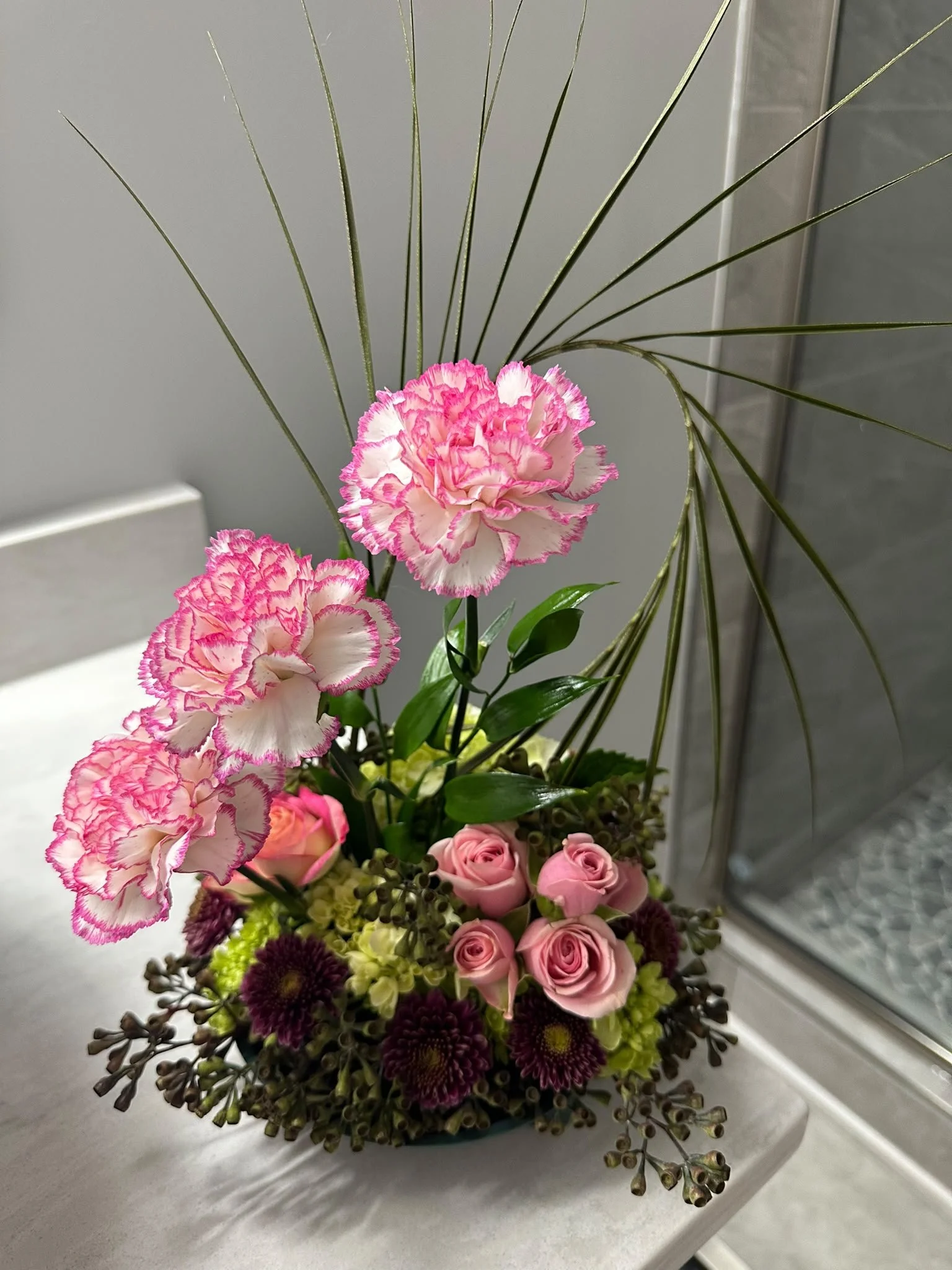 Colorful floral arrangement with pink-edged carnations, pink roses, purple chrysanthemums, and green foliage in a bathroom