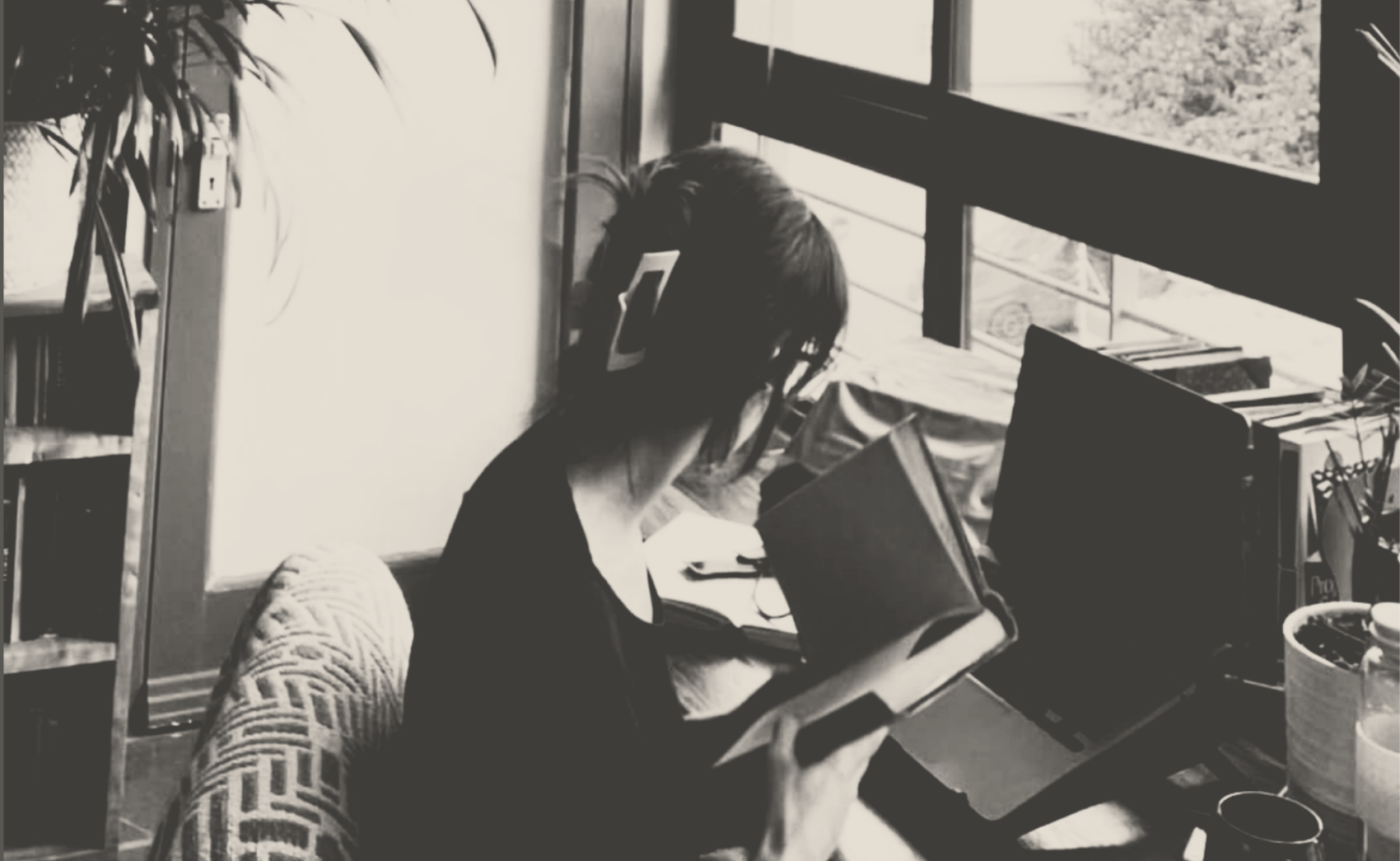 A person sitting at a cluttered desk near a window, reading a book with a tablet, laptop, and various items around.