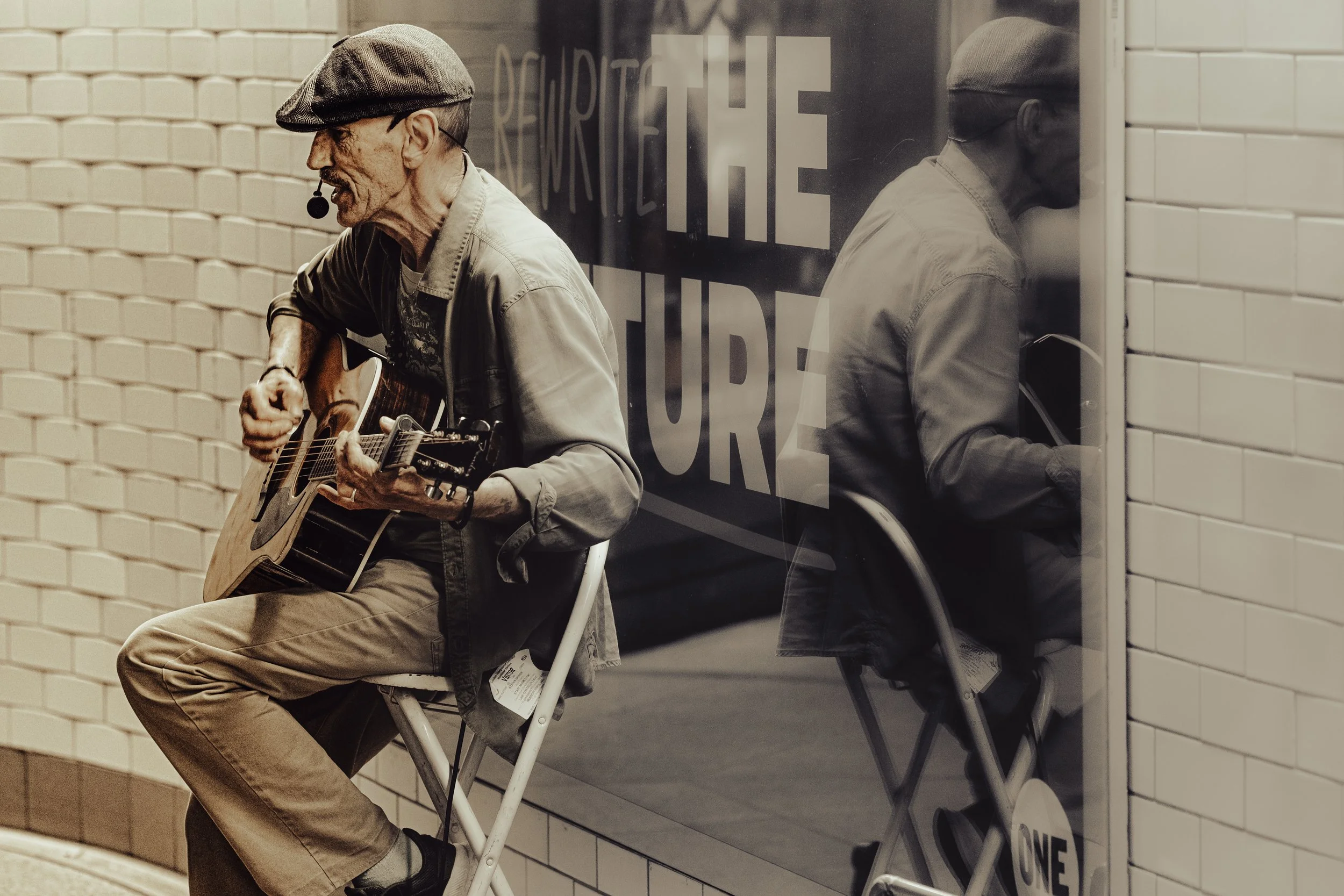 An elderly man with a gray beard and a gray flat cap is sitting on a white folding chair, playing an acoustic guitar and singing into a headset microphone. He is wearing a light-colored jacket and beige pants. The background features white brick wall