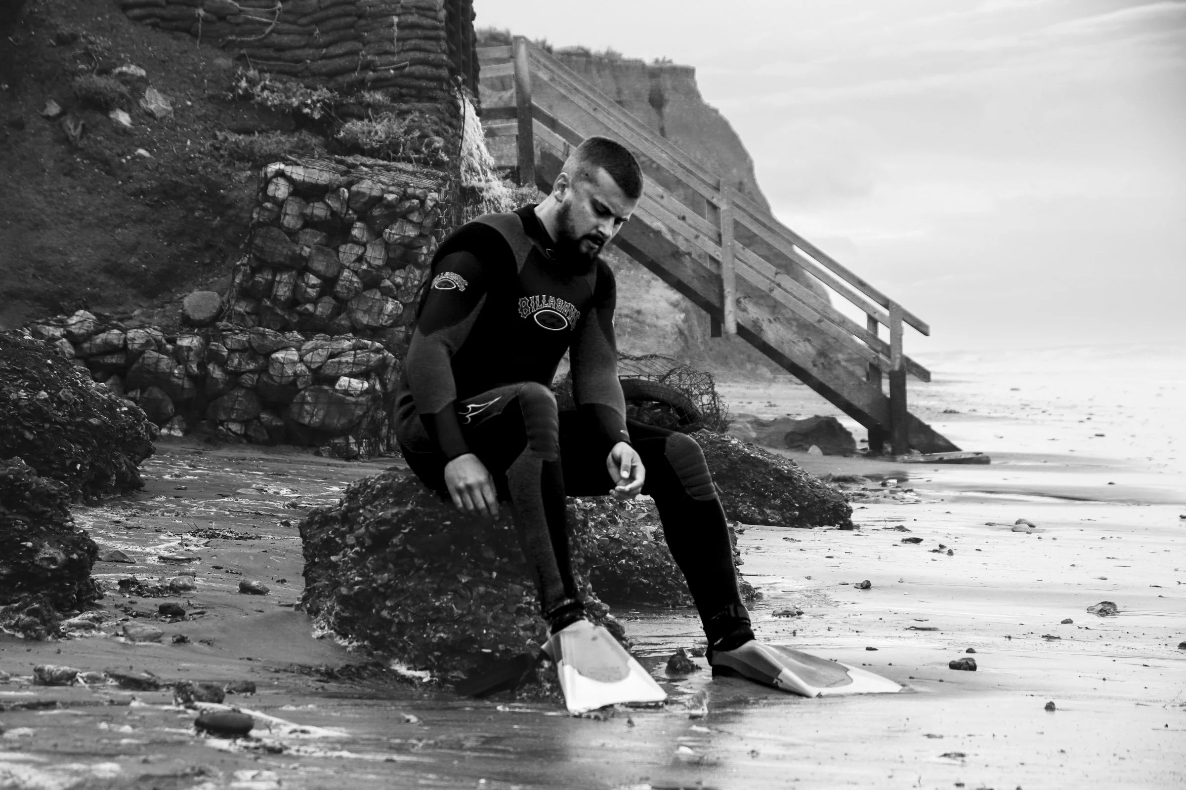 A man sitting on rocks on a beach, looking down, wearing wetsuit and fins, with a wooden staircase and cliff in the background.