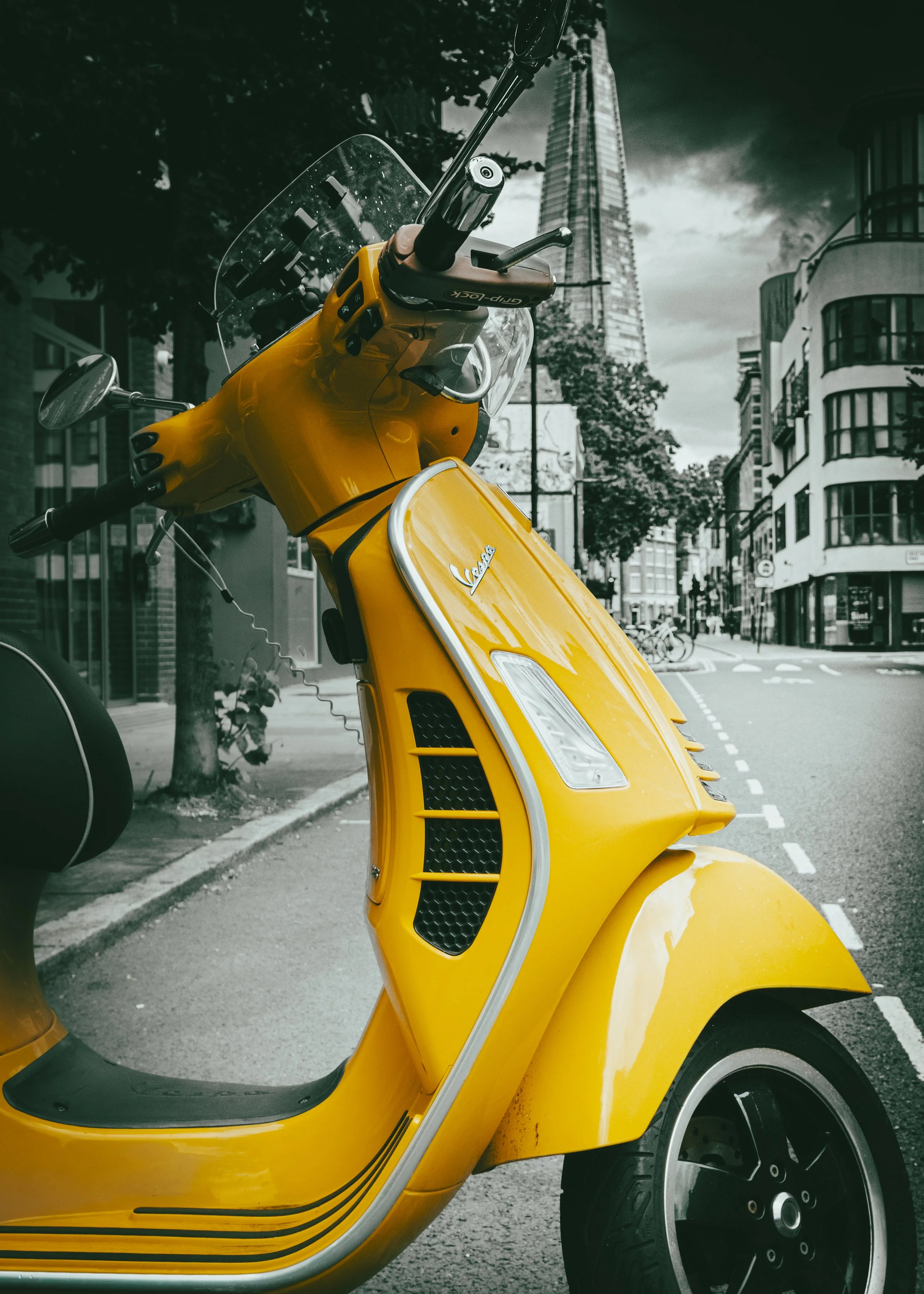 Yellow Vespa scooter parked on city street with tall skyscraper in background, partially in black and white with only the scooter in color.