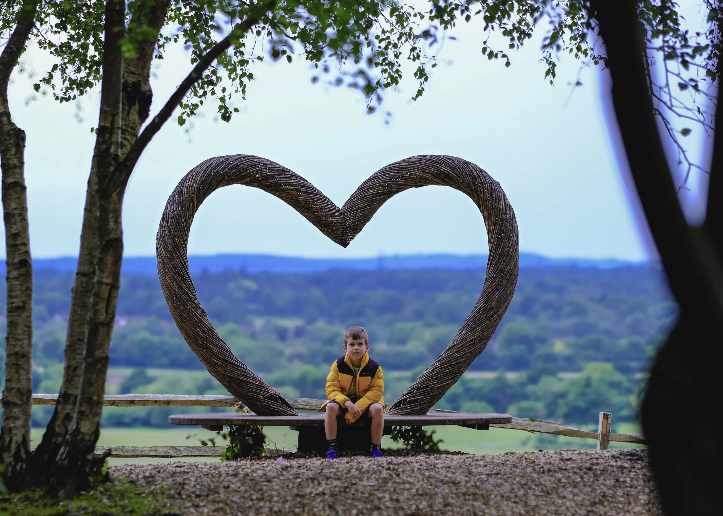 A young boy sitting on a bench in front of a heart-shaped wooden frame with trees and a scenic landscape in the background.