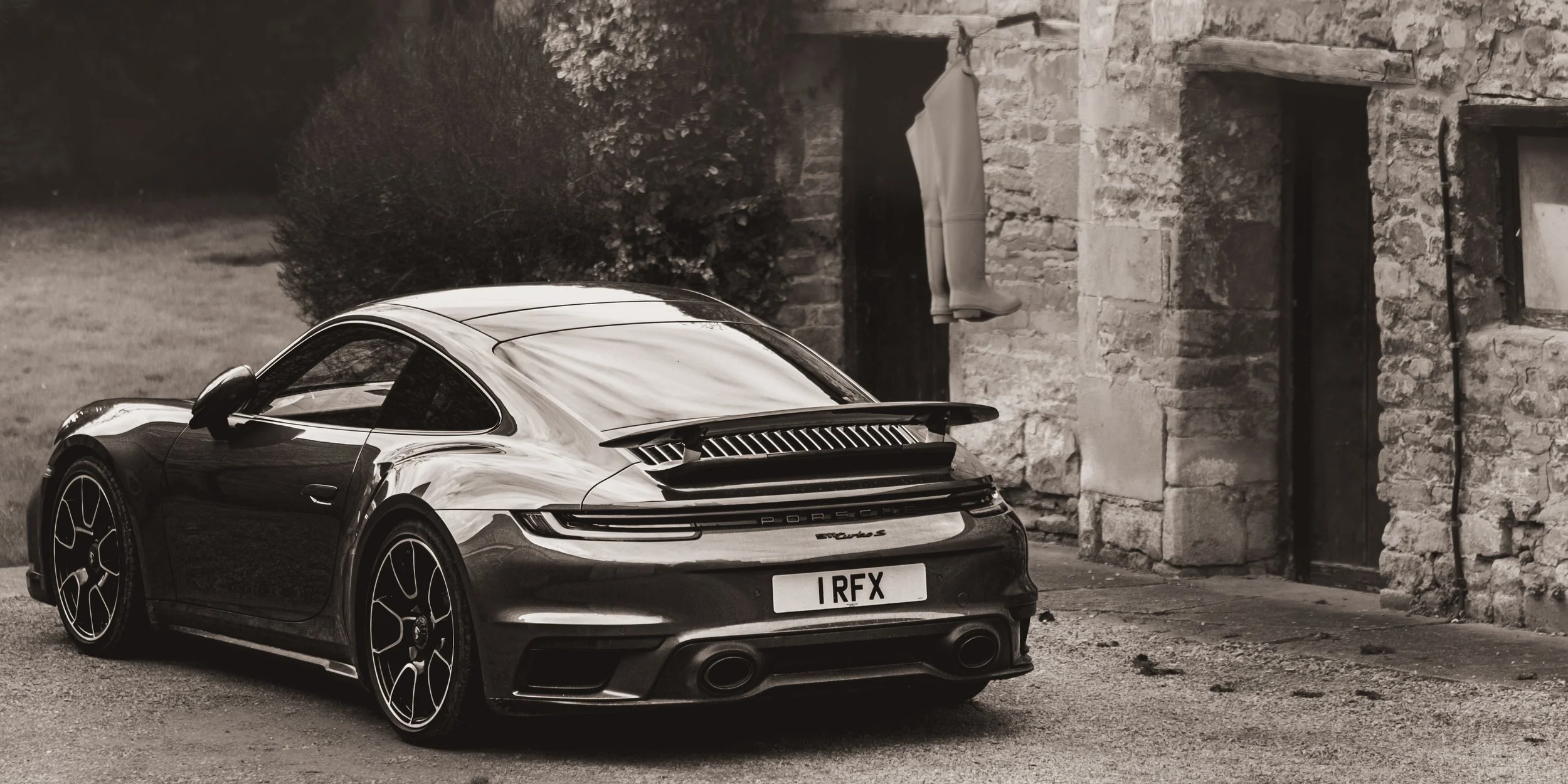 A black Porsche sports car parked outdoors in front of a stone building with a pair of rain boots hanging from a window ledge.