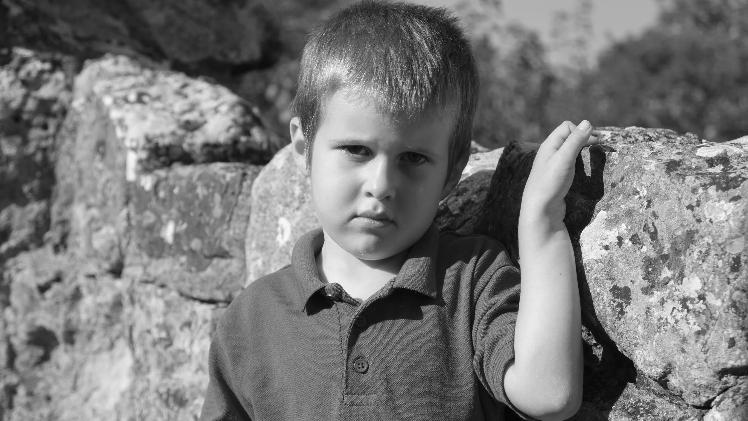 A young boy with short hair wearing a polo shirt, standing beside large rocks outdoors, with a serious expression.
