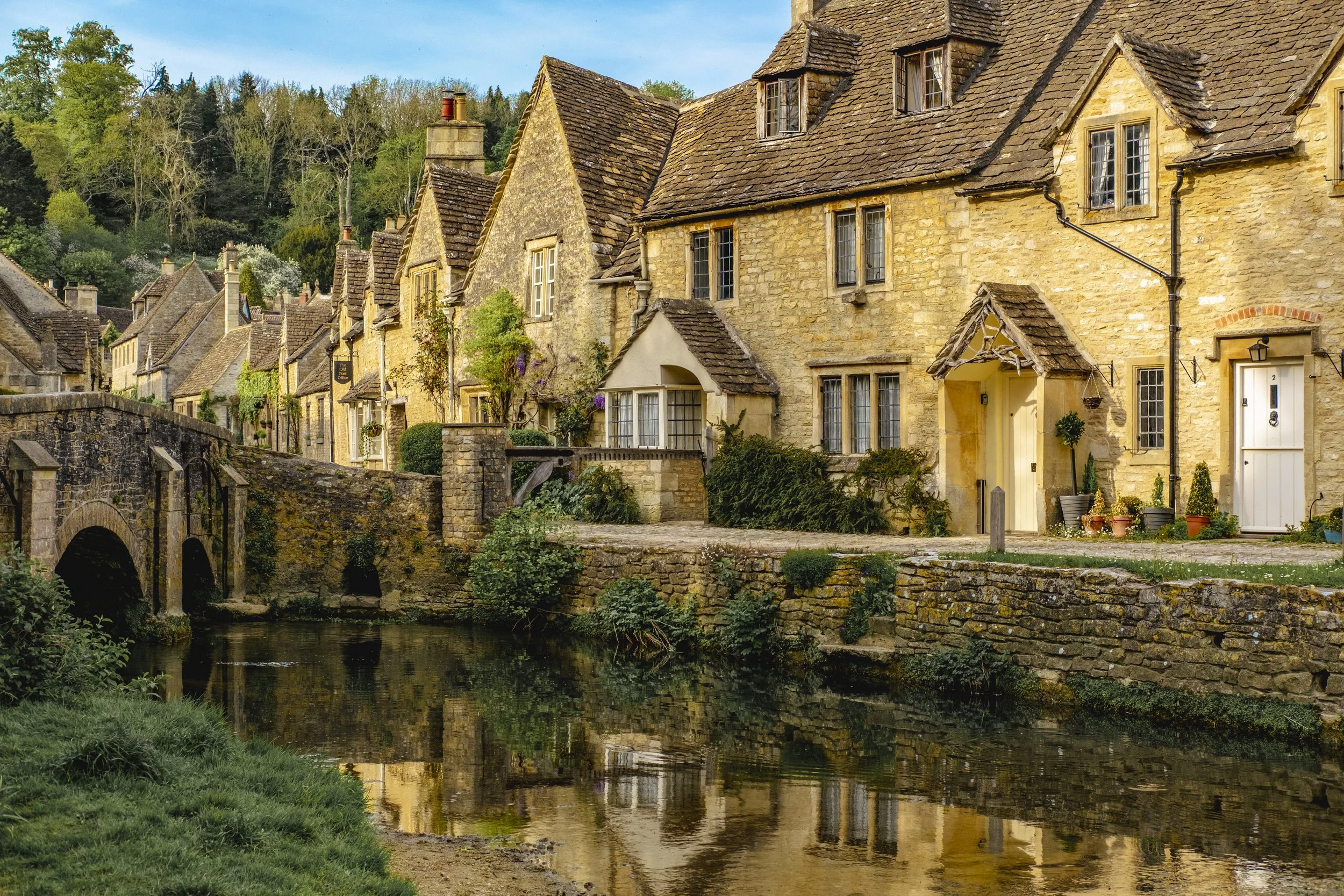 A row of historic stone cottages with steep roofs beside a small river, surrounded by green trees and shrubs.