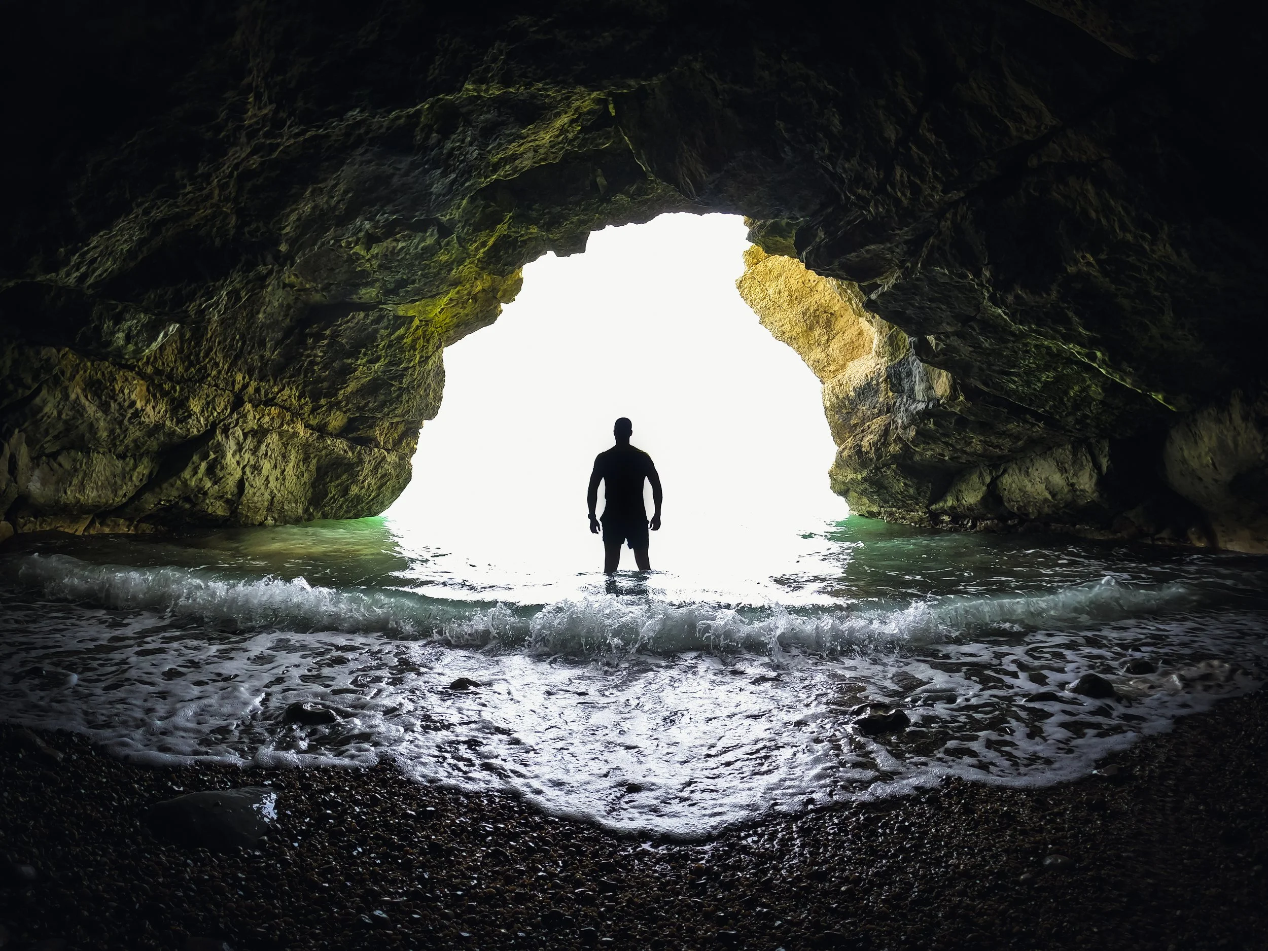 Person standing inside a cave opening looking out towards a bright sky and water, with dark rocky cave walls and a pebble-covered beach in the foreground.