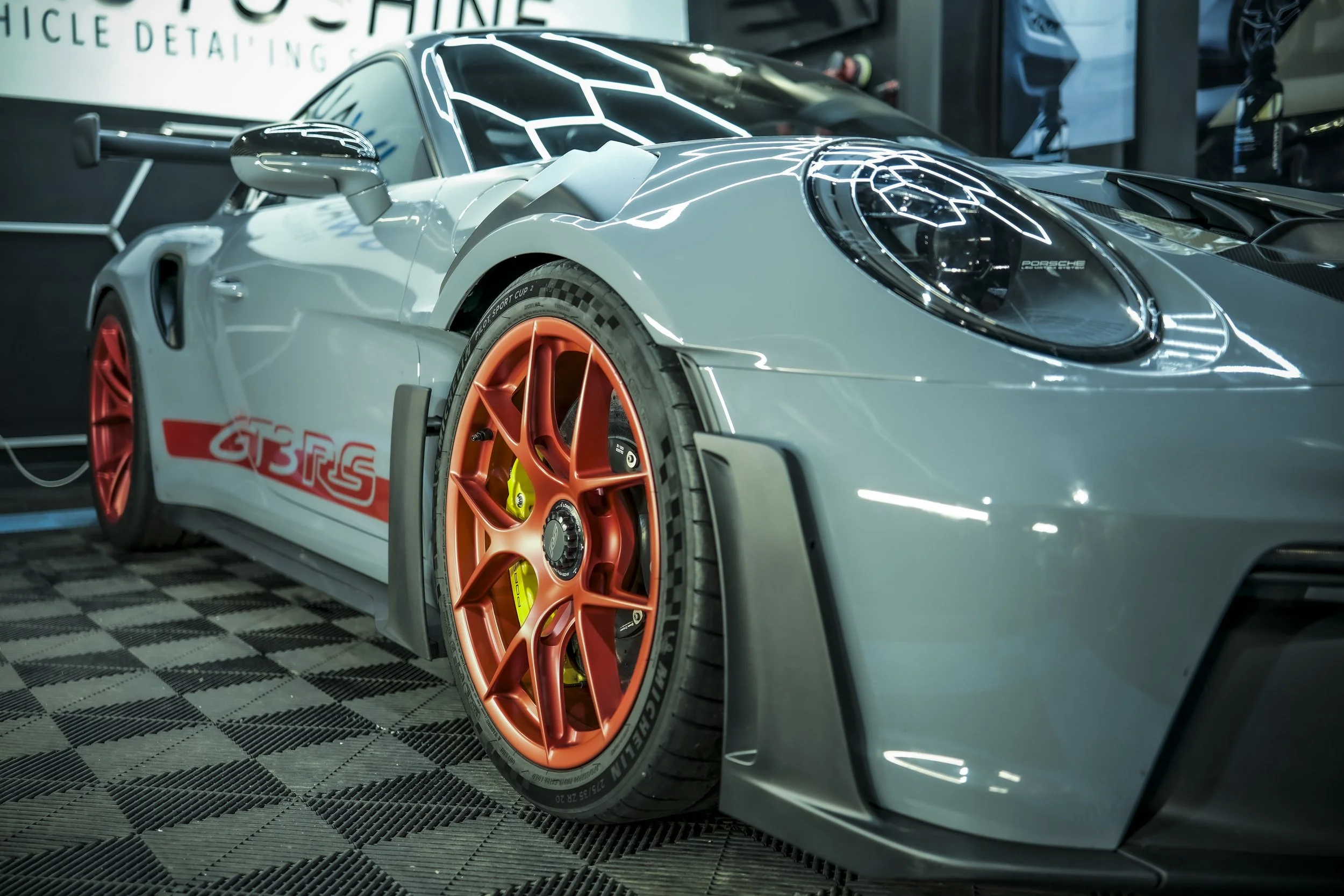 A silver race car with orange wheels and red detailing, parked indoors. The car has a rear wing and aerodynamic features, with a checkered floor underneath.
