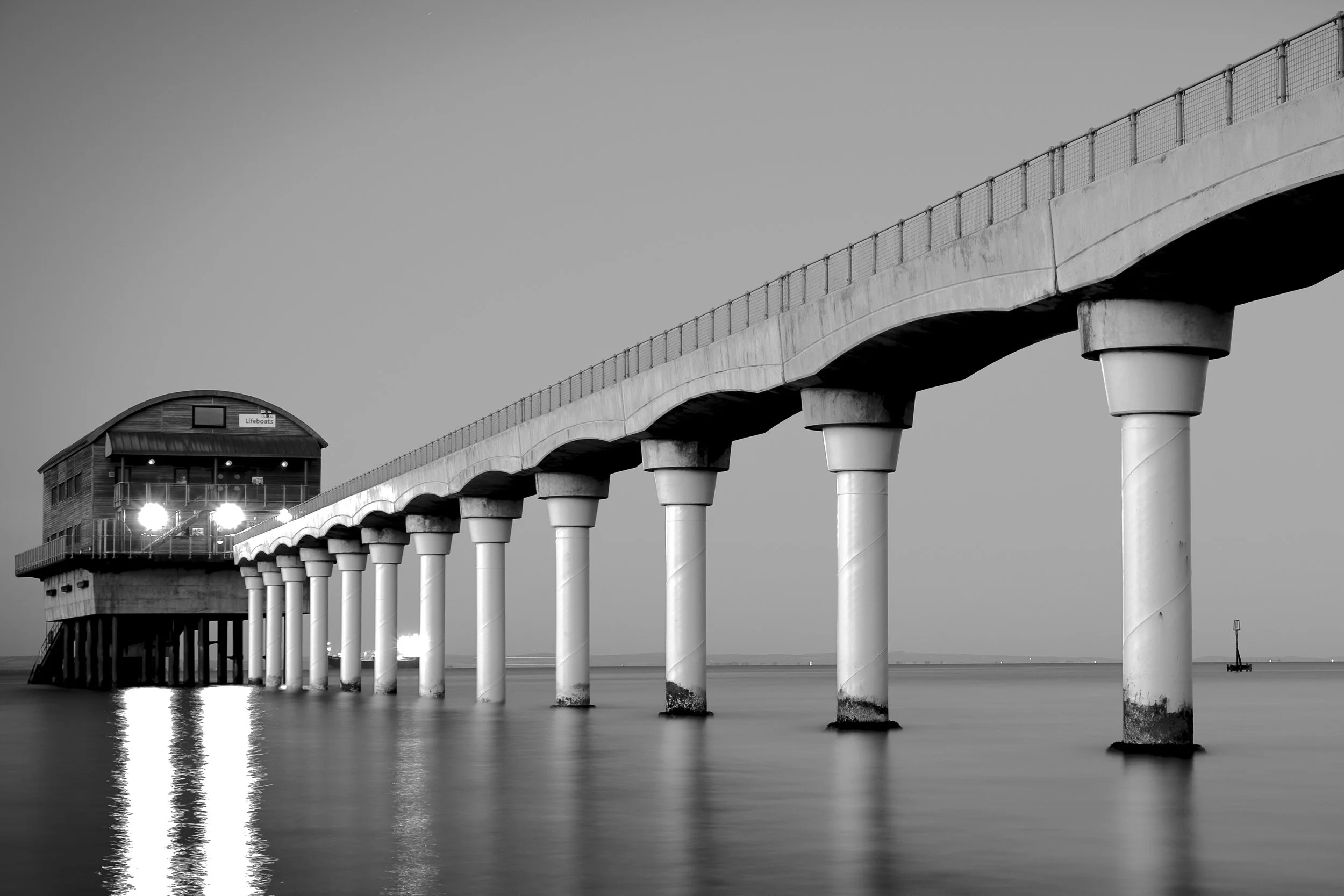 A black and white photo of a pier extending over the water, with a building labeled 'Lifeboats' at the end of the pier and a tiny lighthouse nearby.