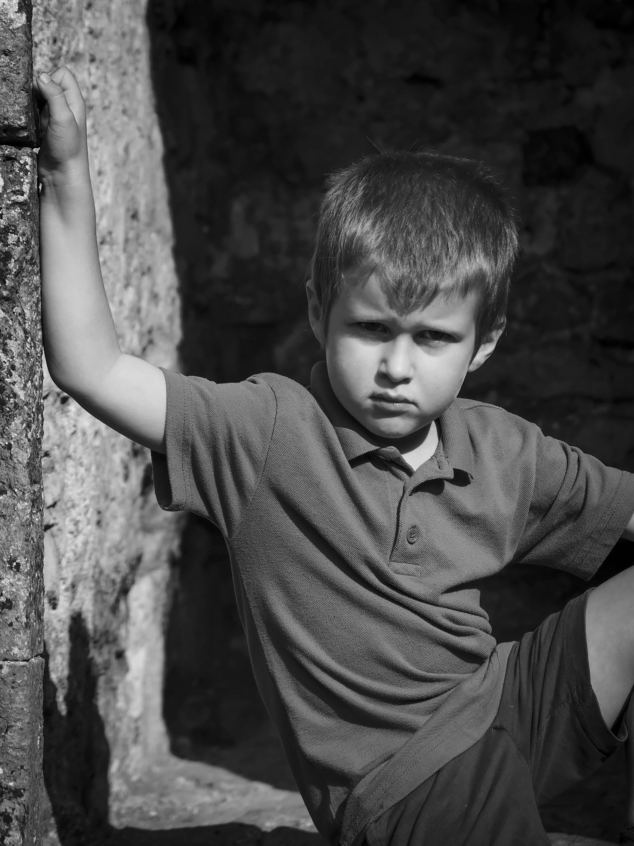 A young boy with short hair wearing a polo shirt, sitting outdoors near a stone wall, with his hand resting against the wall and a serious expression on his face.