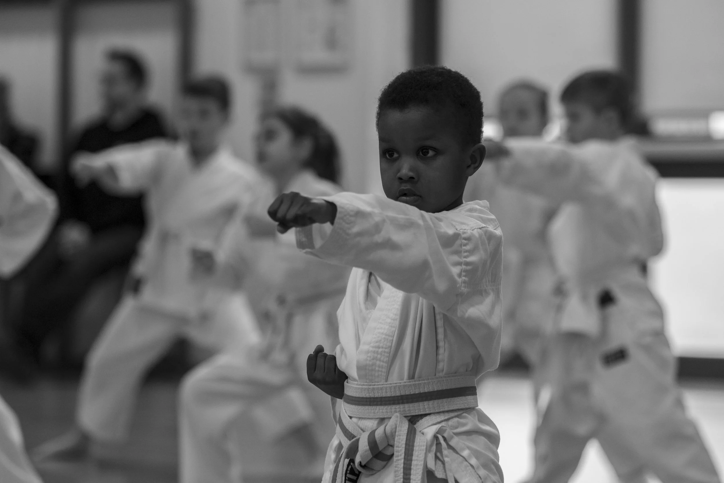 A young boy dressed in a martial arts gi practicing a punch in a dojo with several other students in the background.