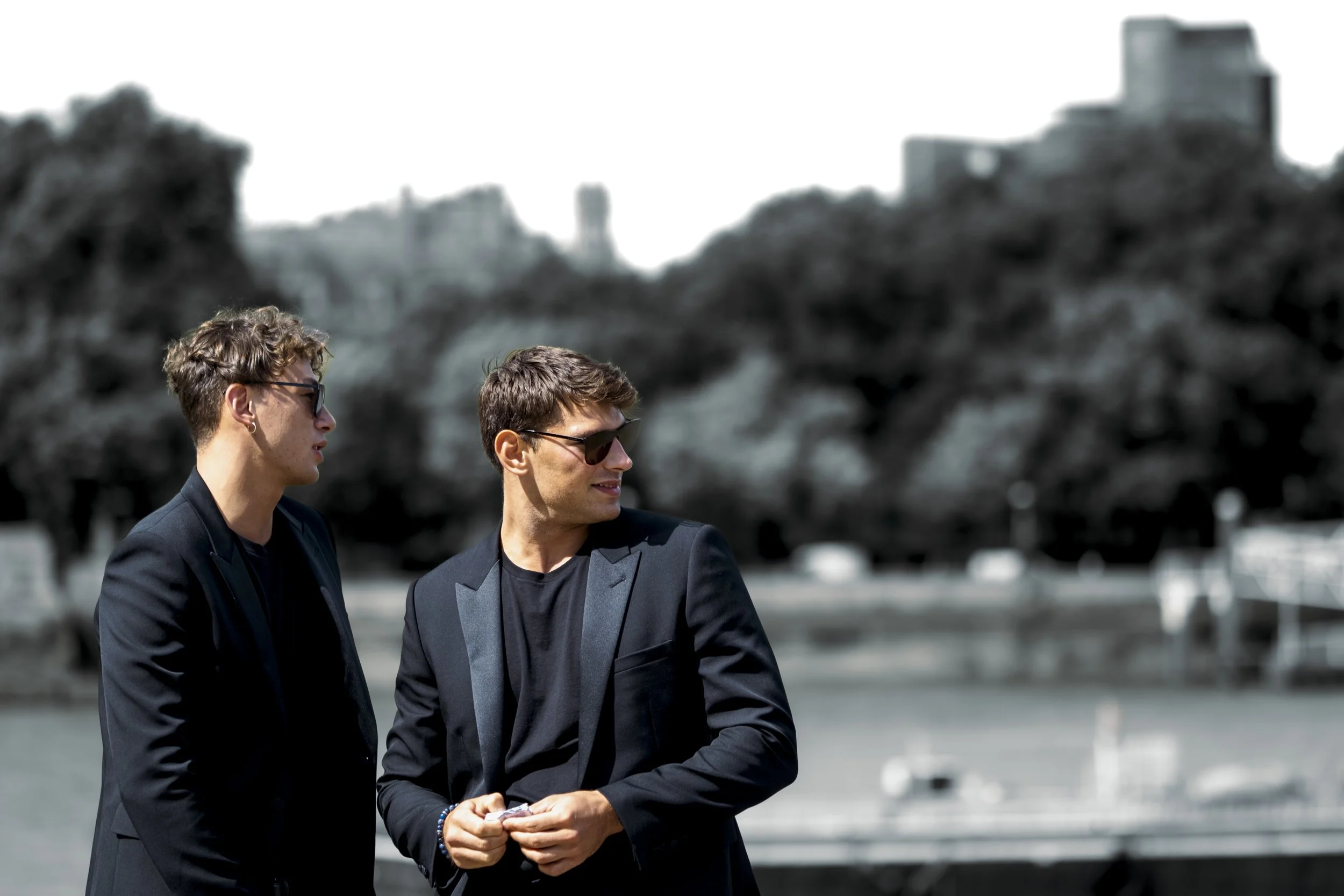 Two young men wearing sunglasses and suits standing outdoors in front of blurred trees and buildings.