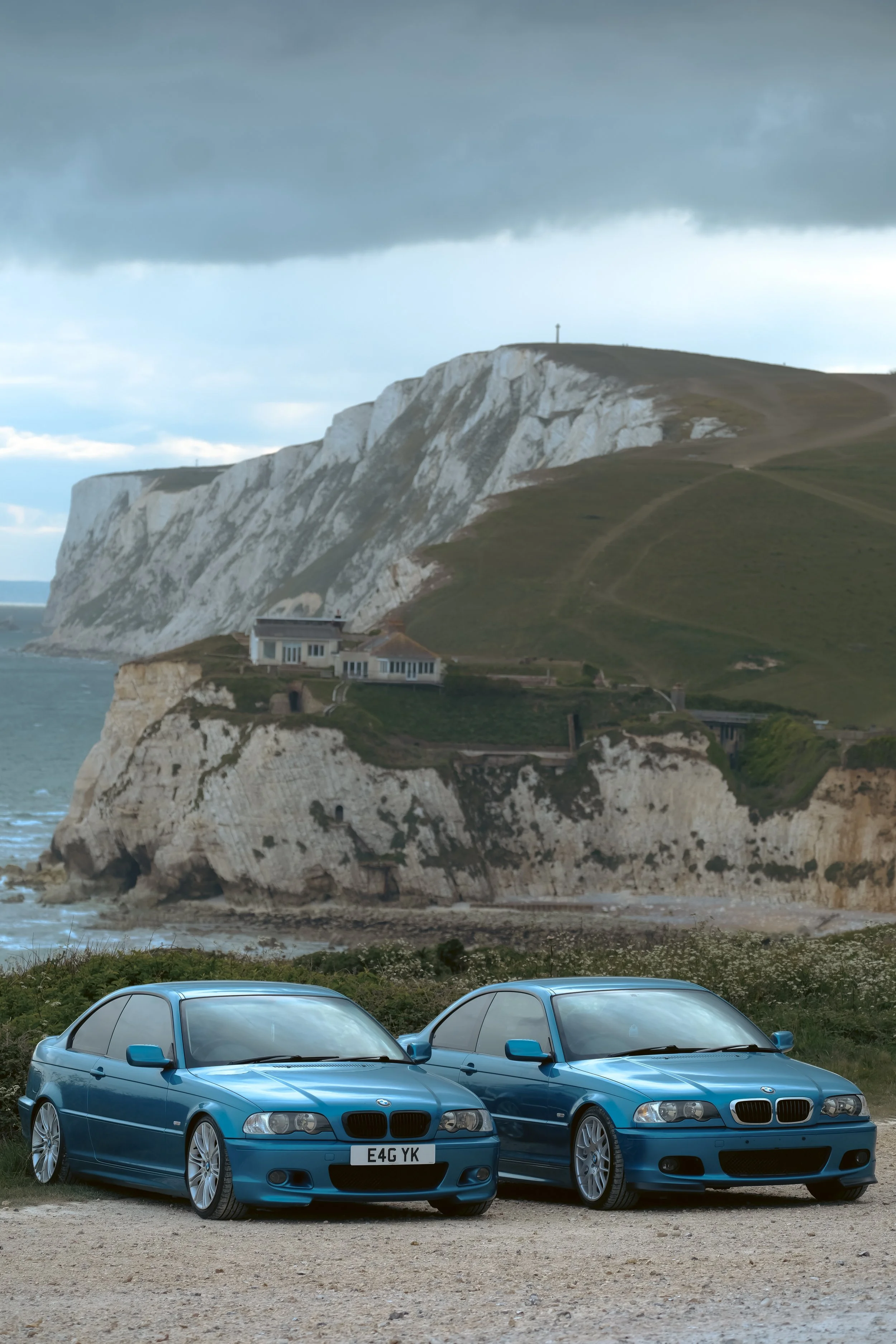 Two blue BMW cars parked on a dirt surface in front of white chalk cliffs with green hills and a cloudy sky in the background.