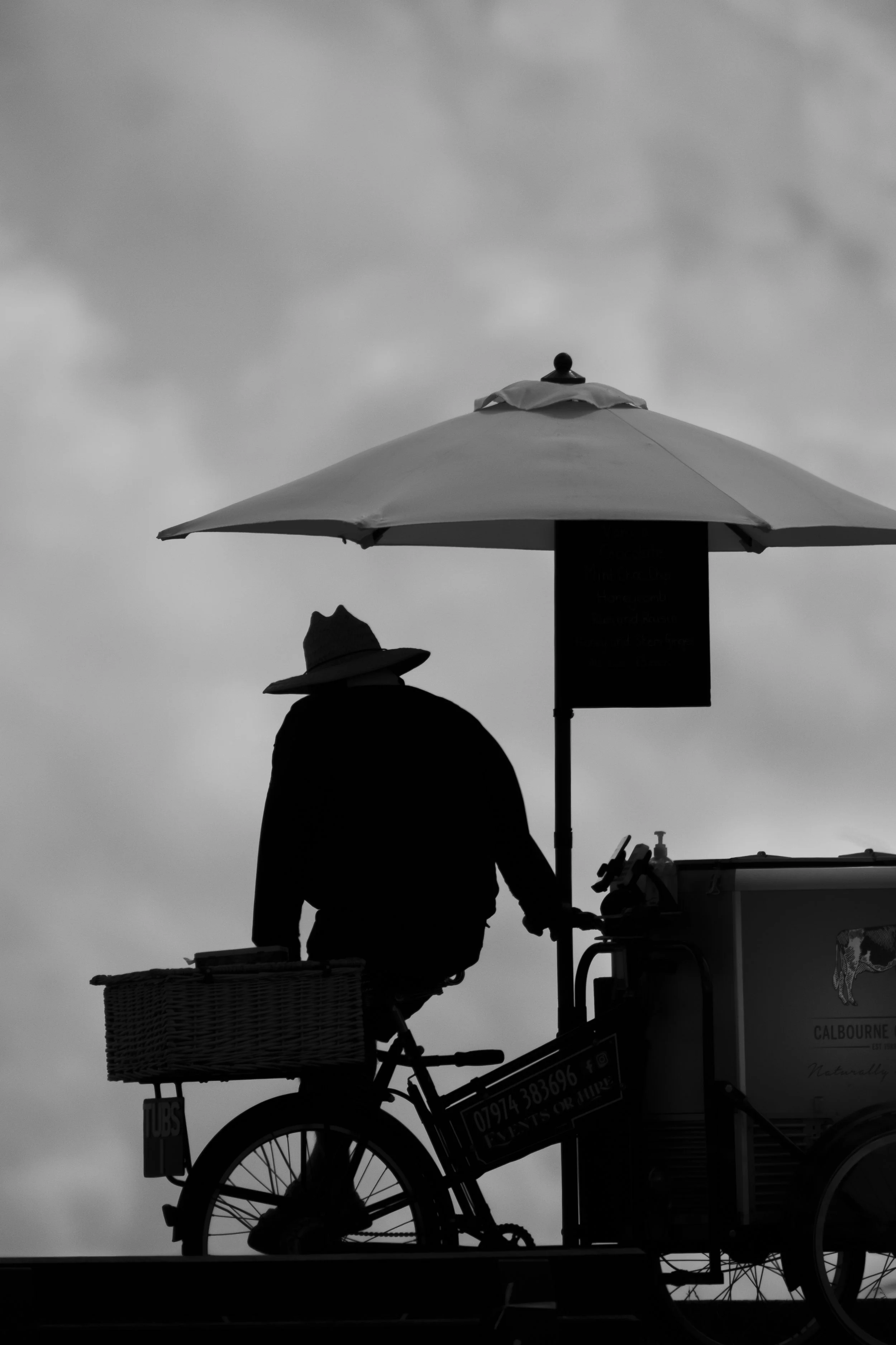 Silhouette of a person riding a bicycle cart with a large umbrella attached, set against a cloudy sky.