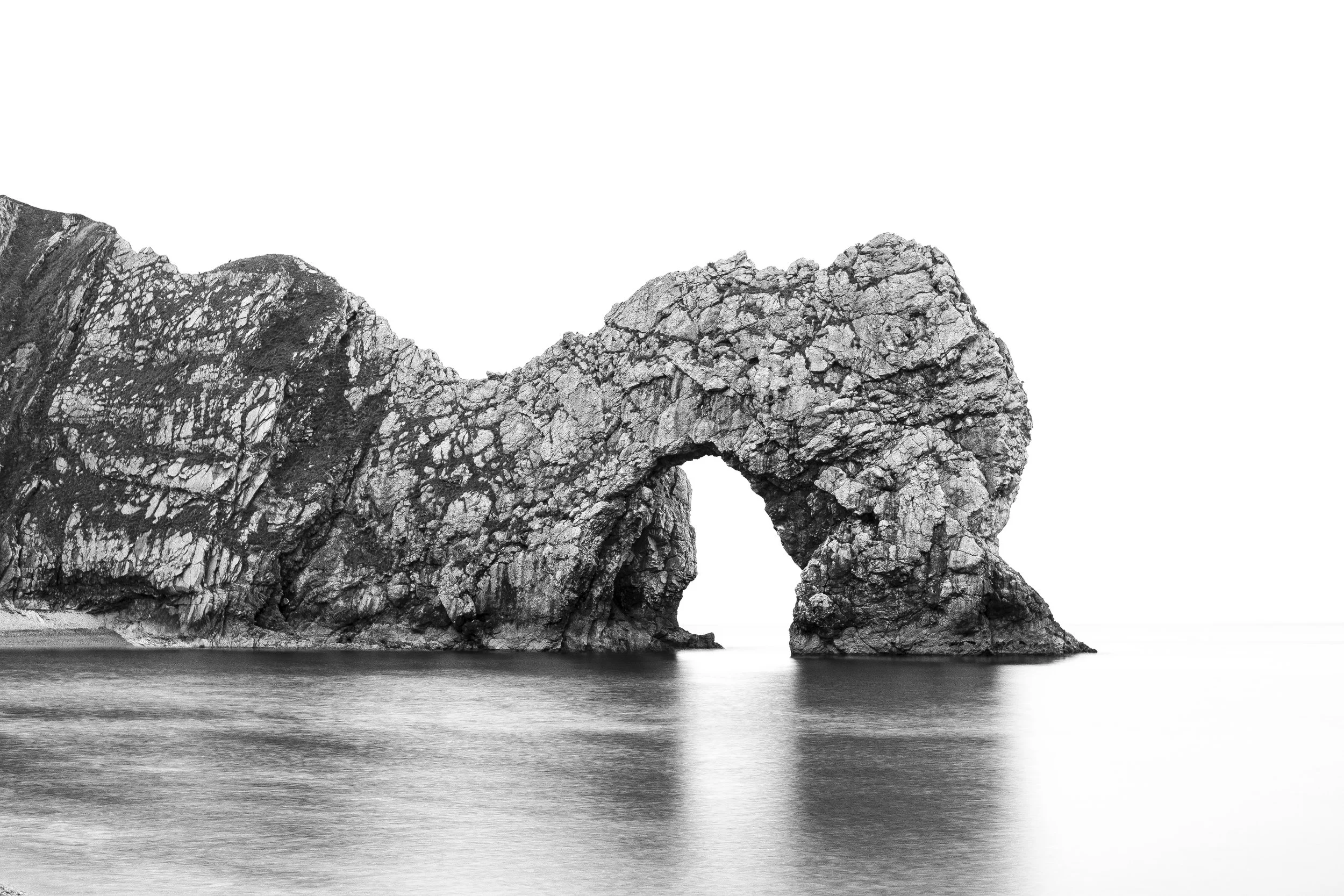 Black and white photo of a natural rock arch formation rising from water, with a clear sky background.